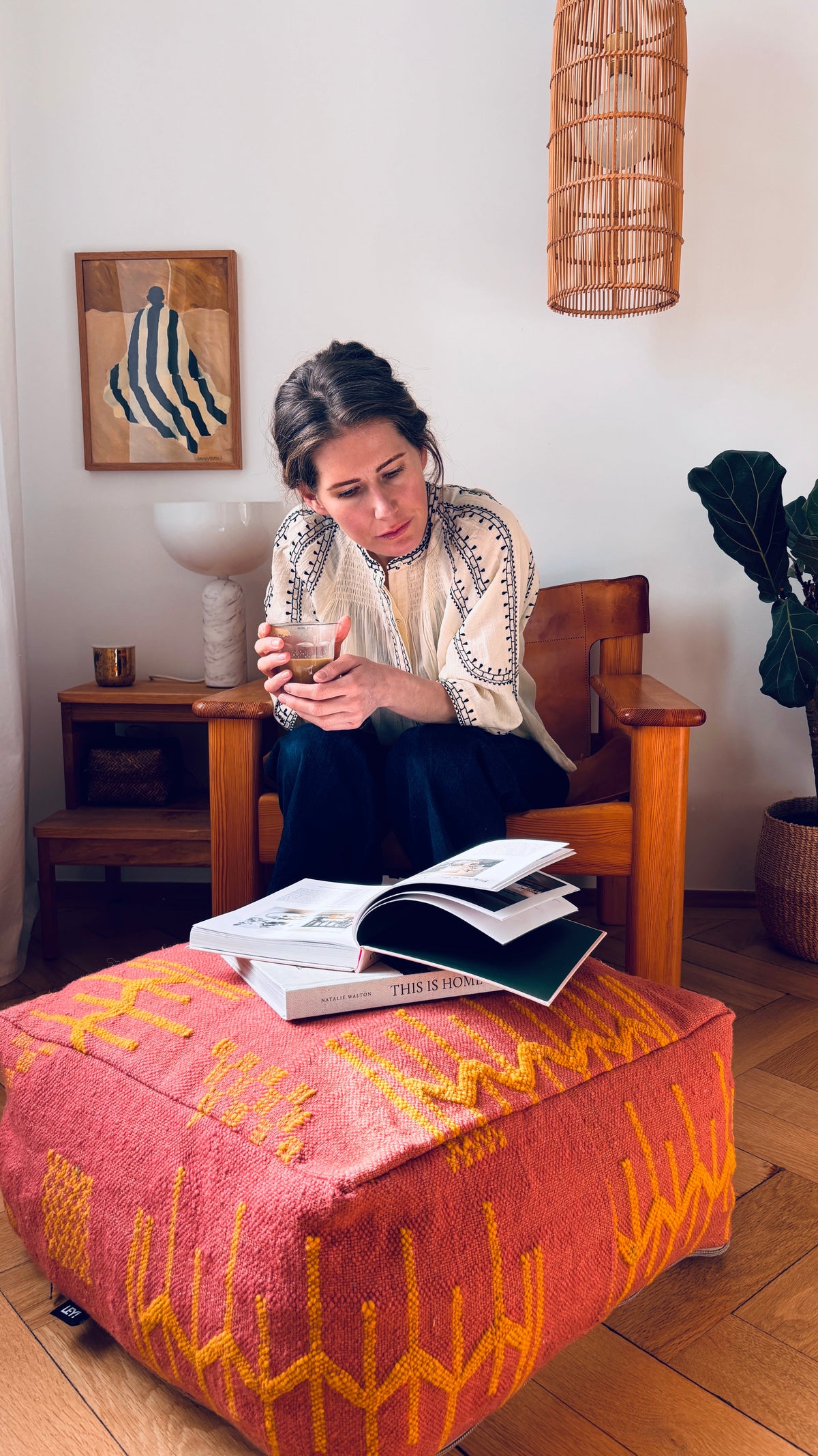 Eine Frau sitzt auf einem Holzstuhl und schaut auf ihr Telefon, während sie Bücher auf einem LEY! Bodenkissen - Colorful Line - Pink Edition.