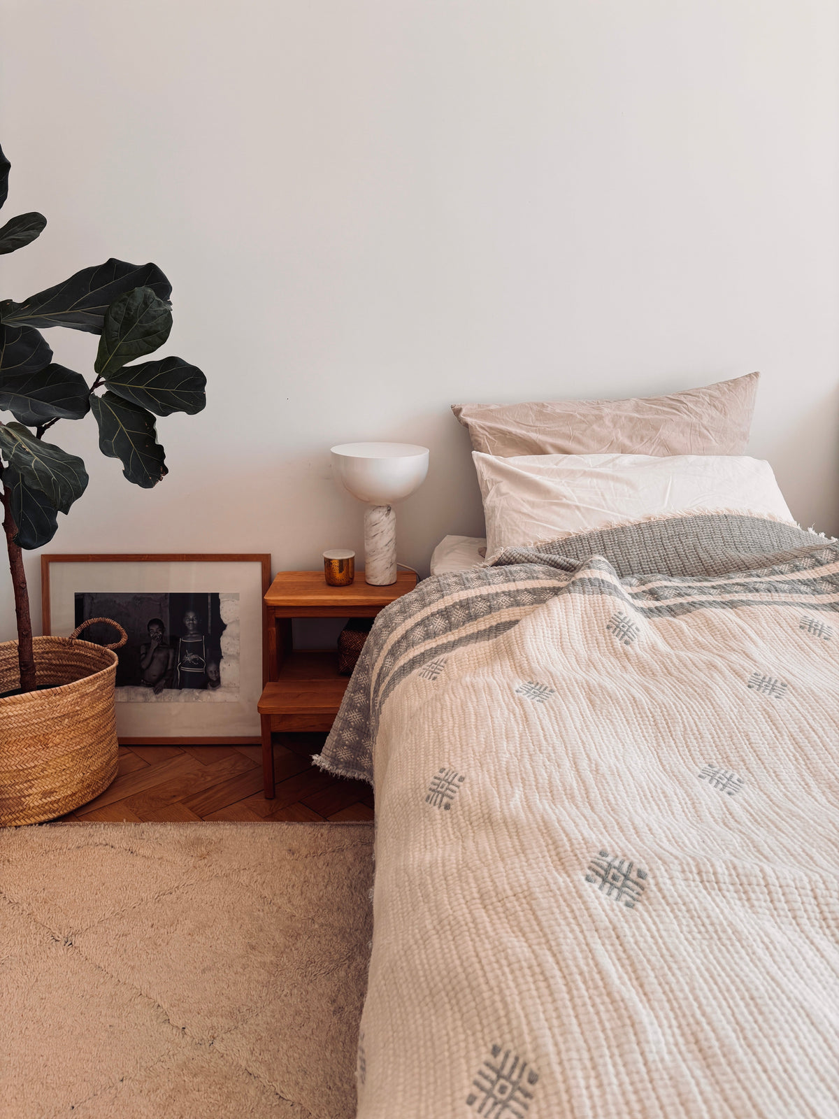 Ein gemütliches Schlafzimmer mit einem ordentlich gemachten Bett mit der LEY! Musselin Decke in Hellgrau-Weiß (2,20 x 2,50 m), beige gemusterter Bettwäsche, Holznachttisch mit weißer Lampe und Kerze, gerahmtem Foto auf dem Boden, geflochtenem Pflanzenkorb und einem Teppich im Boho-Stil.
