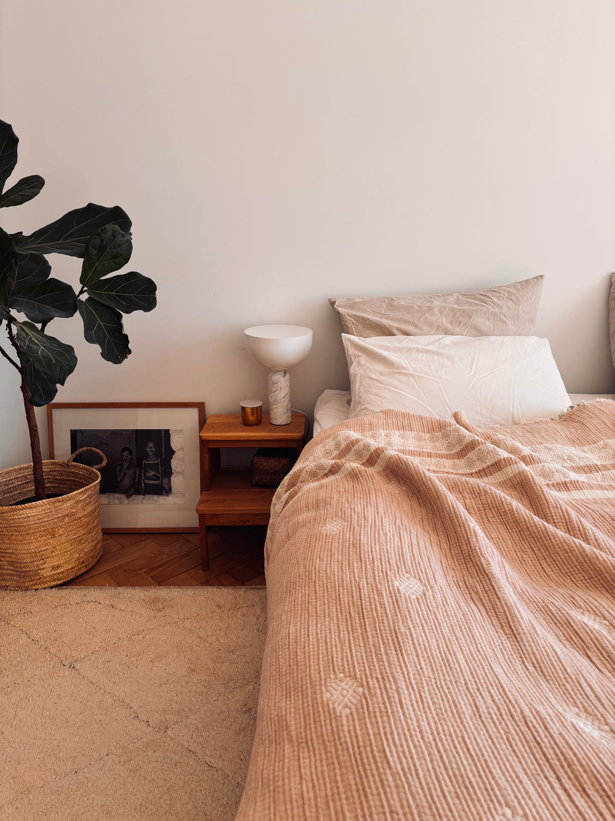 Ein gemütliches Boho-Schlafzimmer mit einem Bett, das mit der LEY! Musselin Decke in Beige-Weiß (2,20 x 2,50 m), weißen Kissen, einem hölzernen Nachttisch mit Lampe und Kerze, einer Topfpflanze und einem gerahmten Foto auf einem hellen Teppichboden.
