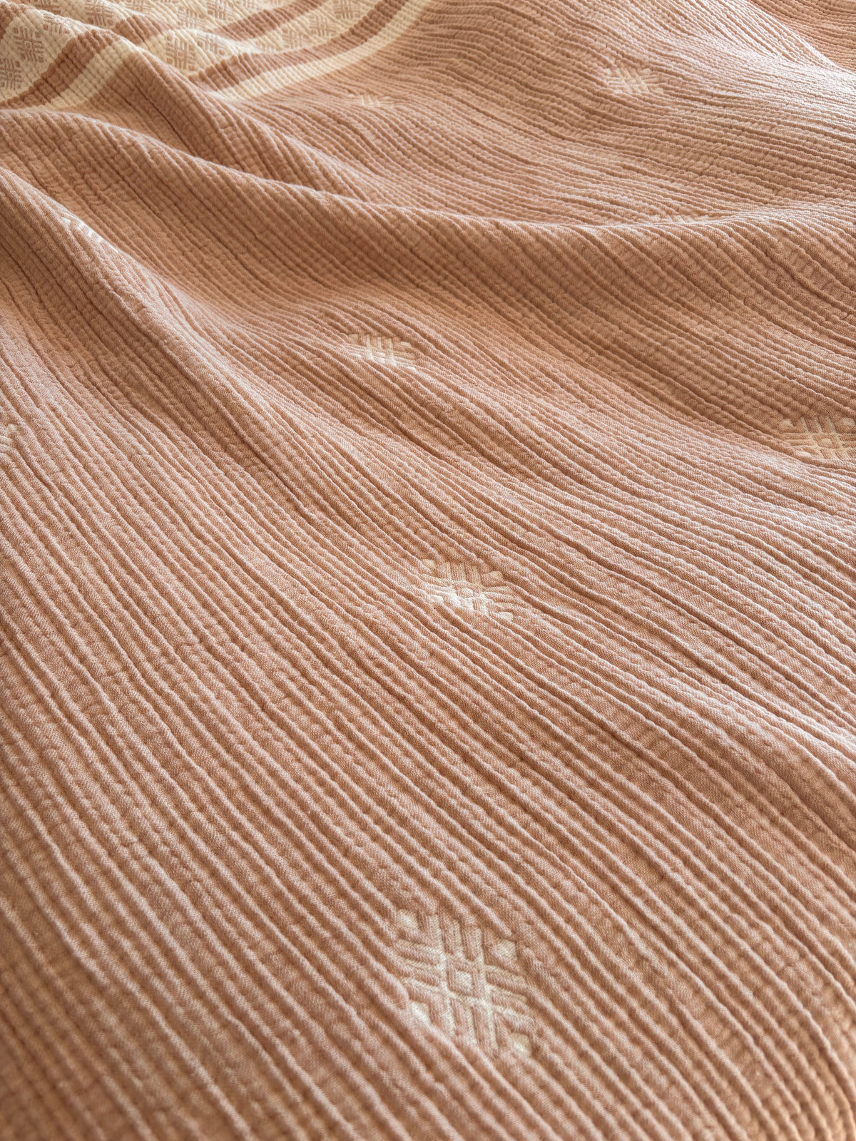 Eine Nahaufnahme der LEY! Musselin Decke in Beige-Weiß (2,20 x 2,50 m), die einen weichen, hellbraunen Boho-Stoff mit weißer, geometrischer Stickerei und feinen vertikalen Linien zeigt, der in sanften Falten drapiert ist.