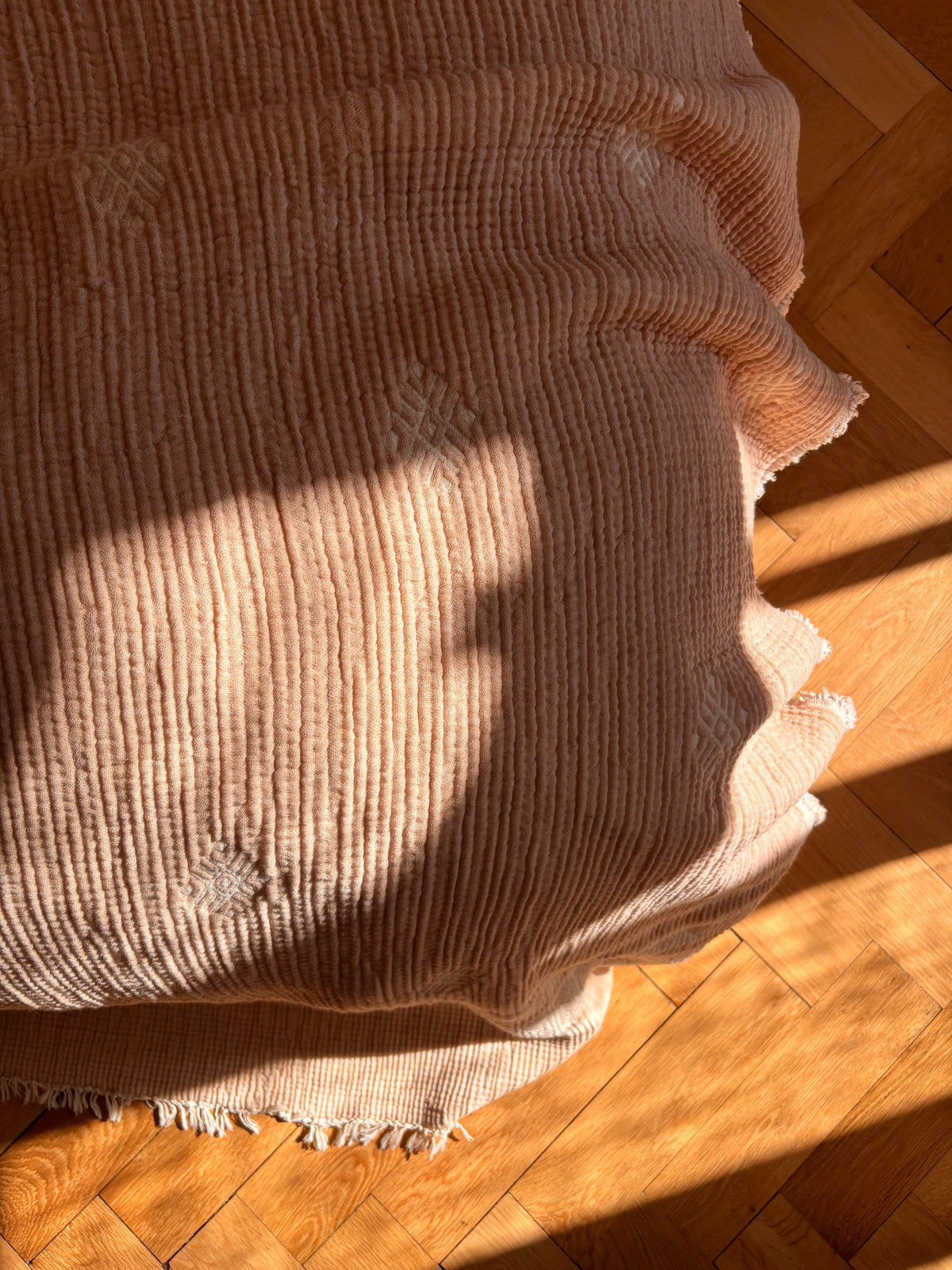 Die Musselin Decke - Beige-Weiß (2,20 x 2,50 m) von LEY! im Boho-Design liegt sanft auf dem Holzboden und wird von warmem Sonnenlicht durchflutet, das die dezenten Muster hervorhebt und jedem Raum einen gemütlichen Charme verleiht.