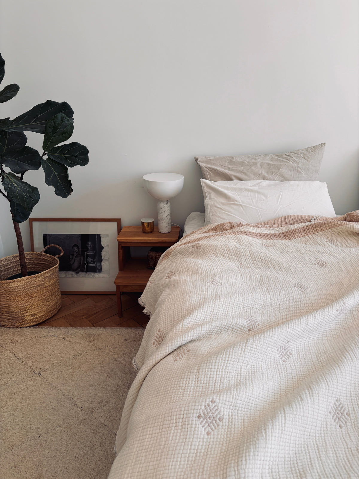 Ein gemütliches Schlafzimmer mit einem hübsch gemachten Bett, das mit der LEY! Musselin Decke in Beige-Weiß (2,20 x 2,50 m), einem hölzernen Nachttisch mit einer weißen Lampe und einer Kerze, einer Topfpflanze, einem gerahmten Kunstwerk, Boho-Akzenten und einem geflochtenen Korb auf einem hellen Teppich.