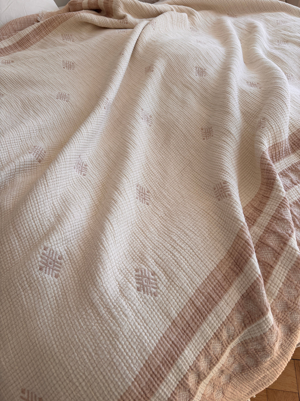 Die LEY! Musselin Decke in Beige-Weiß (2,20 x 2,50 m) zeigt ein dezentes geometrisches Muster und gestreifte Bordüren, drapiert über einem Bett für einen gemütlichen Boho-Look mit weichen Falten.