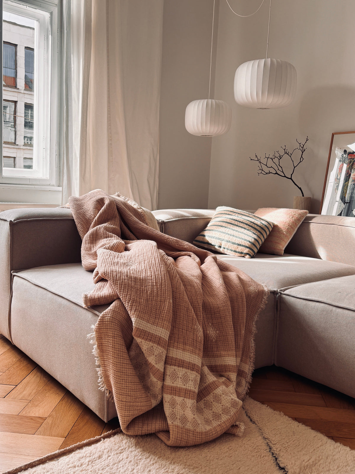 In einer gemütlichen Wohnzimmerecke steht ein modernes beiges Sektionssofa, drapiert mit der LEY! Musselin Decke in Beige-Weiß (2,20 x 2,50 m), gepaart mit einem gestreiften Kopfkissen. Drei runde Pendelleuchten hängen darüber, während das Sonnenlicht durch das Fenster fällt.