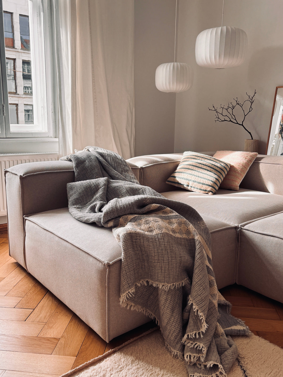 Ein gemütliches, modernes Wohnzimmer mit einer taupefarbenen Couchgarnitur, gestreiften Kissen, LEY! Musselin Decke in Hellgrau-Weiß (2,20 x 2,50 m), zwei weiße Pendelleuchten, ein dekorativer Ast in einer Vase und ein großes Fenster für natürliches Boho-Design-Flair.