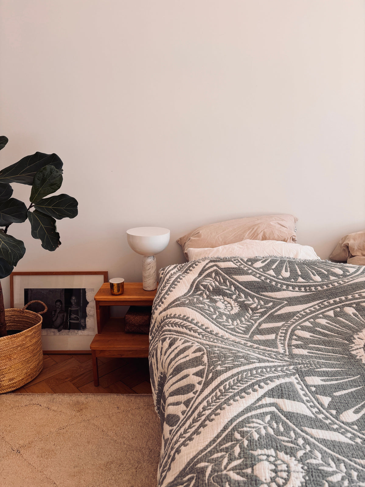 In einer gemütlichen Schlafecke steht eine LEY! CALMA Musselin Decke in Dunkelgrau (2,20 x 2,30 m) auf dem Bett, mit einem hölzernen Nachttisch, einer Lampe, einer Tasse, einer Topfpflanze, einem Weidenkorb, einem Bilderrahmen und einem beigen Teppich auf dem Holzboden.