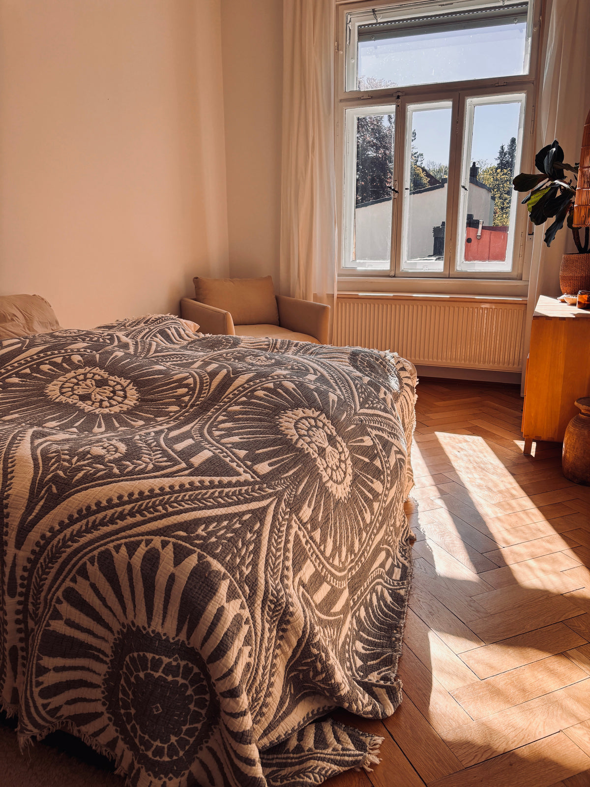 In einem gemütlichen, sonnendurchfluteten Schlafzimmer im Boho-Stil liegt die CALMA Musselin Decke in Dunkelgrau (2,20 x 2,30 m) von LEY! drapiert auf dem Bett, mit einem Sessel, einer Pflanze und einer Kommode am Fenster, und Sonnenlicht auf dem Holzboden.
