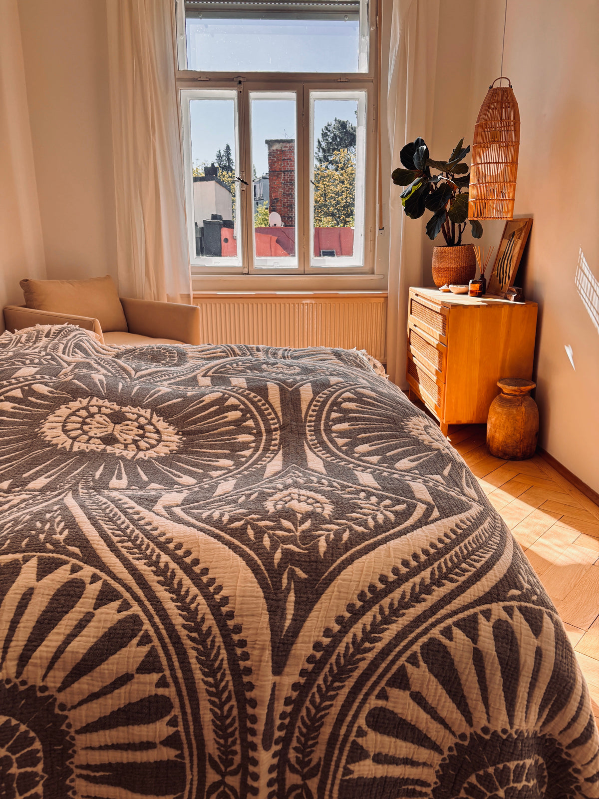 Ein gemütliches Schlafzimmer mit Sonnenlicht, das mit der CALMA Musselin Decke - Dunkelgrau (2,20 x 2,30 m) von LEY! im Boho-Stil, einer Holzkommode mit Dekoration, einer Topfpflanze und neutral getönten Wänden und Möbeln ausgestattet ist.