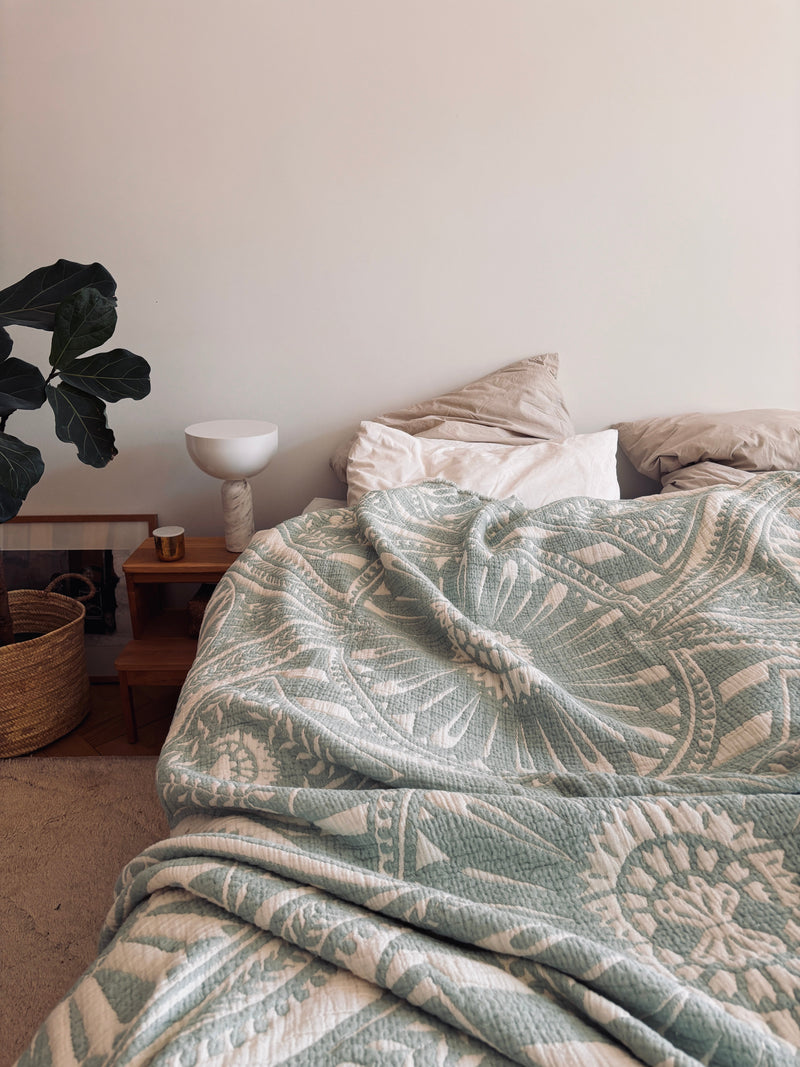 Ein gemütliches, ungemachtes Bett mit der CALMA Musselin Decke von LEY! in mint (2,20 x 2,30 m), gemusterter grün-weißer Bettwäsche, neutralen Kissen und einem hölzernen Nachttisch mit Dekor schafft echten Boho-Design-Charme.