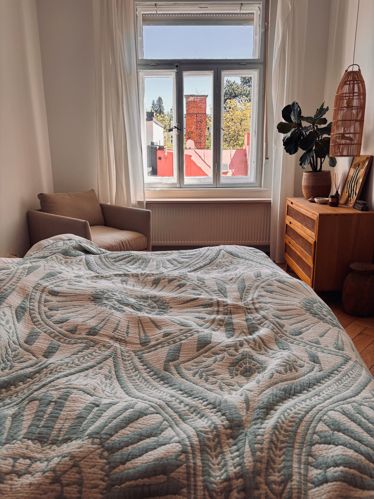 Ein helles Schlafzimmer mit einer LEY! CALMA Musselin Decke in Mint (2,20 x 2,30 m) auf dem Bett, einem beigen Sessel am Fenster, einer hölzernen Kommode, einer Topfpflanze und Sonnenlicht, das durch die Fenster strömt und die roten und weißen Gebäude draußen zeigt.
