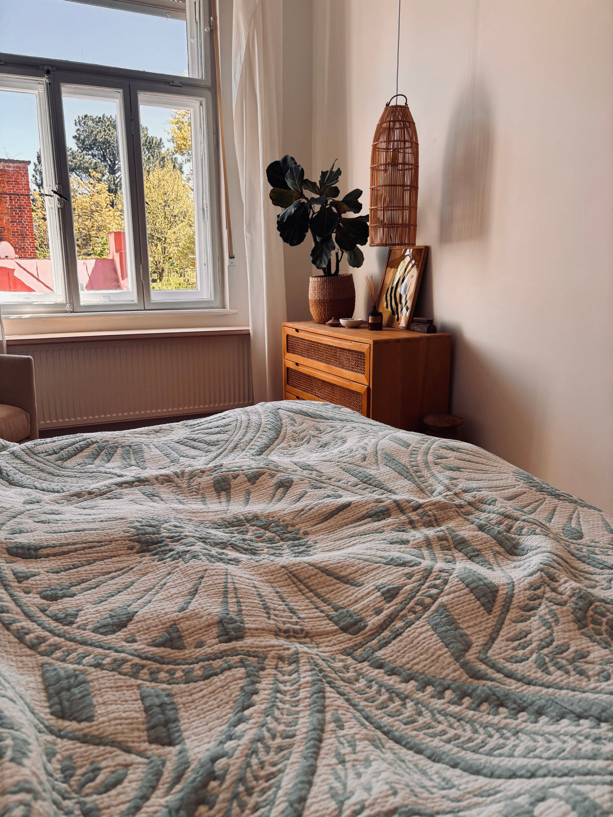Ein gemütliches Schlafzimmer mit der LEY! CALMA Musselin Decke in Mint (2,20 x 2,30 m) auf dem Bett, eine Holzkommode mit Topfpflanzen und Boho-Akzenten, eine Pendellampe aus Korbgeflecht und Sonnenlicht, das an Bäumen und roten Dächern vorbeifließt.