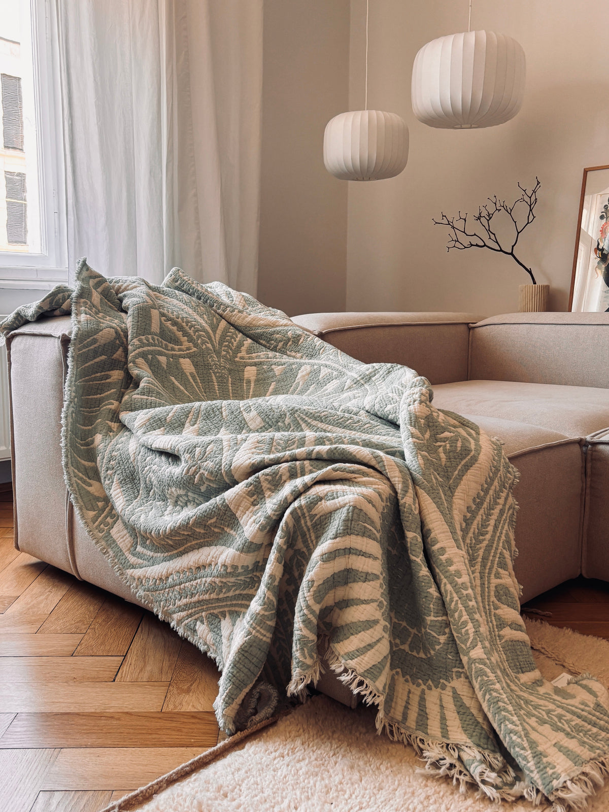 Die CALMA Musselin Decke von LEY! in Mint (2,20 x 2,30 m) wird in einem gemütlichen Wohnzimmer mit Holzboden und modernen weißen Pendelleuchten über einem hellen Sofa drapiert und verleiht dem Raum einen luftigen, stilvollen Touch.