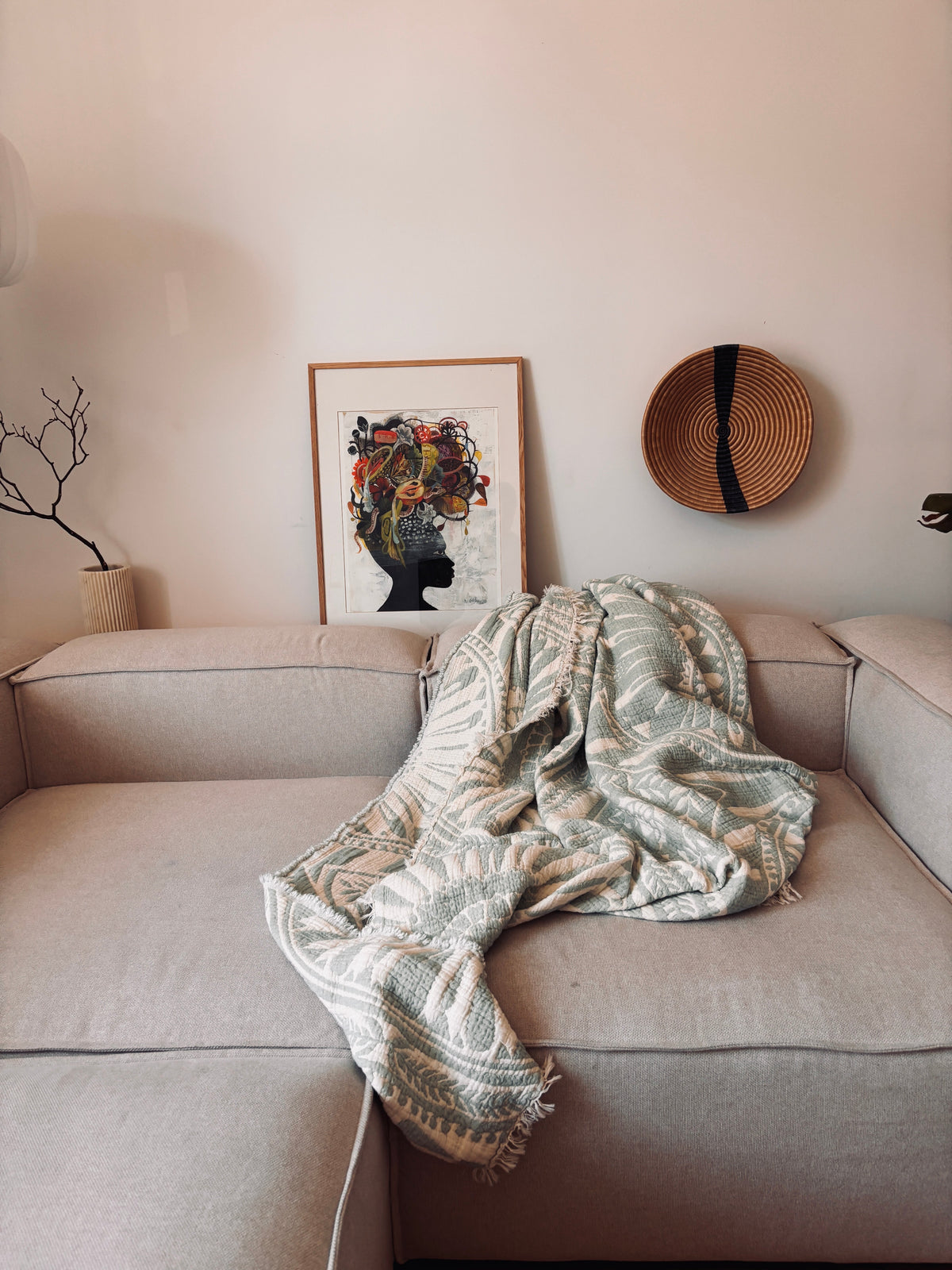 Ein hellbeiges Sofa wird mit der LEY! CALMA Musselin Decke in Mint (2,20 x 2,30 m) drapiert, um einen luftigen, atmungsaktiven Look zu erzielen. Dahinter vervollständigen Boho-Akzente wie gerahmte abstrakte Kunst und ein runder geflochtener Korb die Szene; eine Vase mit Zweigen steht auf der linken Armlehne.