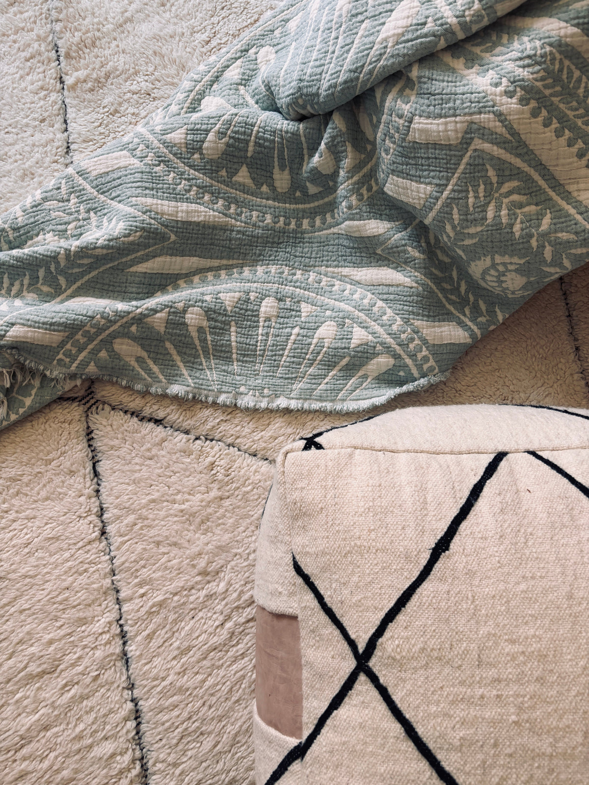 Die CALMA Musselin Decke in Mint (2,20 x 2,30 m) von LEY! liegt teilweise auf einem cremefarbenen Strukturteppich mit schwarzen Gitterlinien. Rechts unten ist ein Kissen mit passendem Boho-Muster und einem beigen Aufnäher zu sehen.