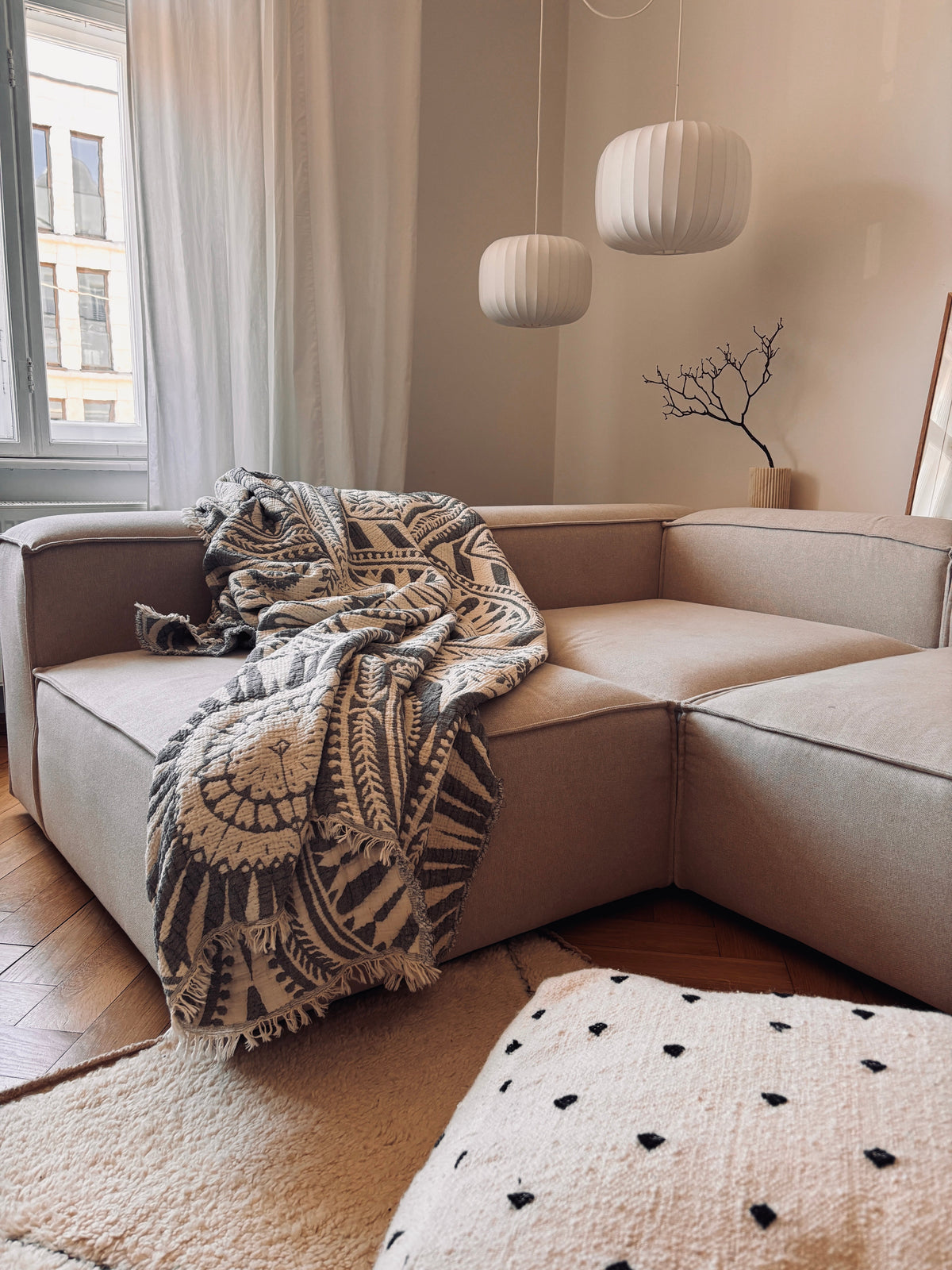 Ein gemütliches Wohnzimmer mit einem beigen Sektionssofa, der CALMA Musselin Decke in Dunkelgrau (LEY!, 2,20 x 2,30 m), modernen weißen Pendelleuchten, einem dekorativen Zweig in einer Vase und transparenten Vorhängen, die das natürliche Licht hereinlassen.