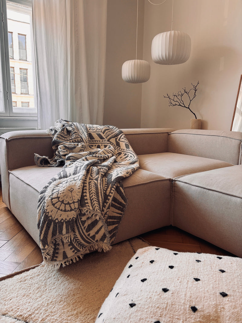 Ein gemütliches Wohnzimmer mit einem beigen Sektionssofa, der CALMA Musselin Decke in Dunkelgrau (LEY!, 2,20 x 2,30 m), modernen weißen Pendelleuchten, einem dekorativen Zweig in einer Vase und transparenten Vorhängen, die das natürliche Licht hereinlassen.