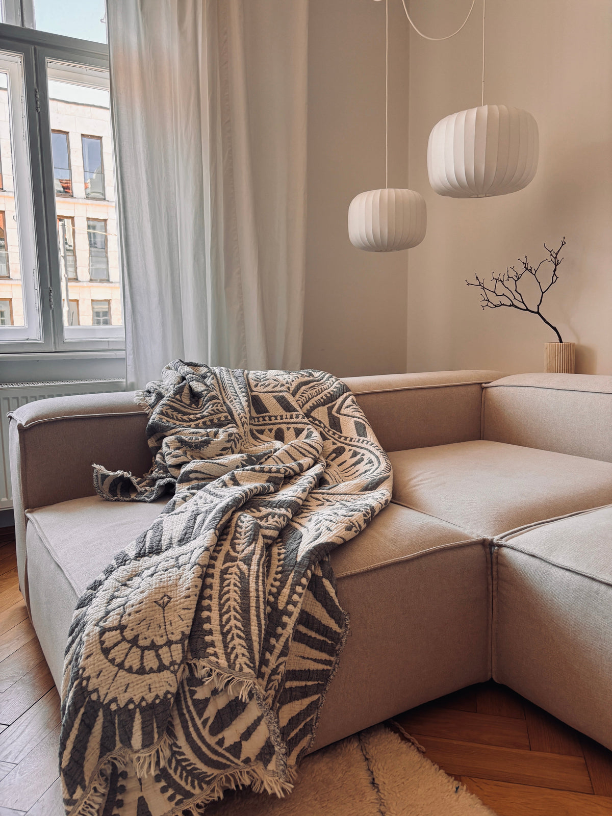 Ein gemütliches beigefarbenes Sektionssofa, über das die CALMA Musselin Decke in Dunkelgrau von LEY! drapiert ist, neben einem großen Fenster mit transparenten Vorhängen. Darüber hängen drei weiße Pendelleuchten, und eine minimalistische Vase mit Zweigen unterstreicht das Boho-Design.