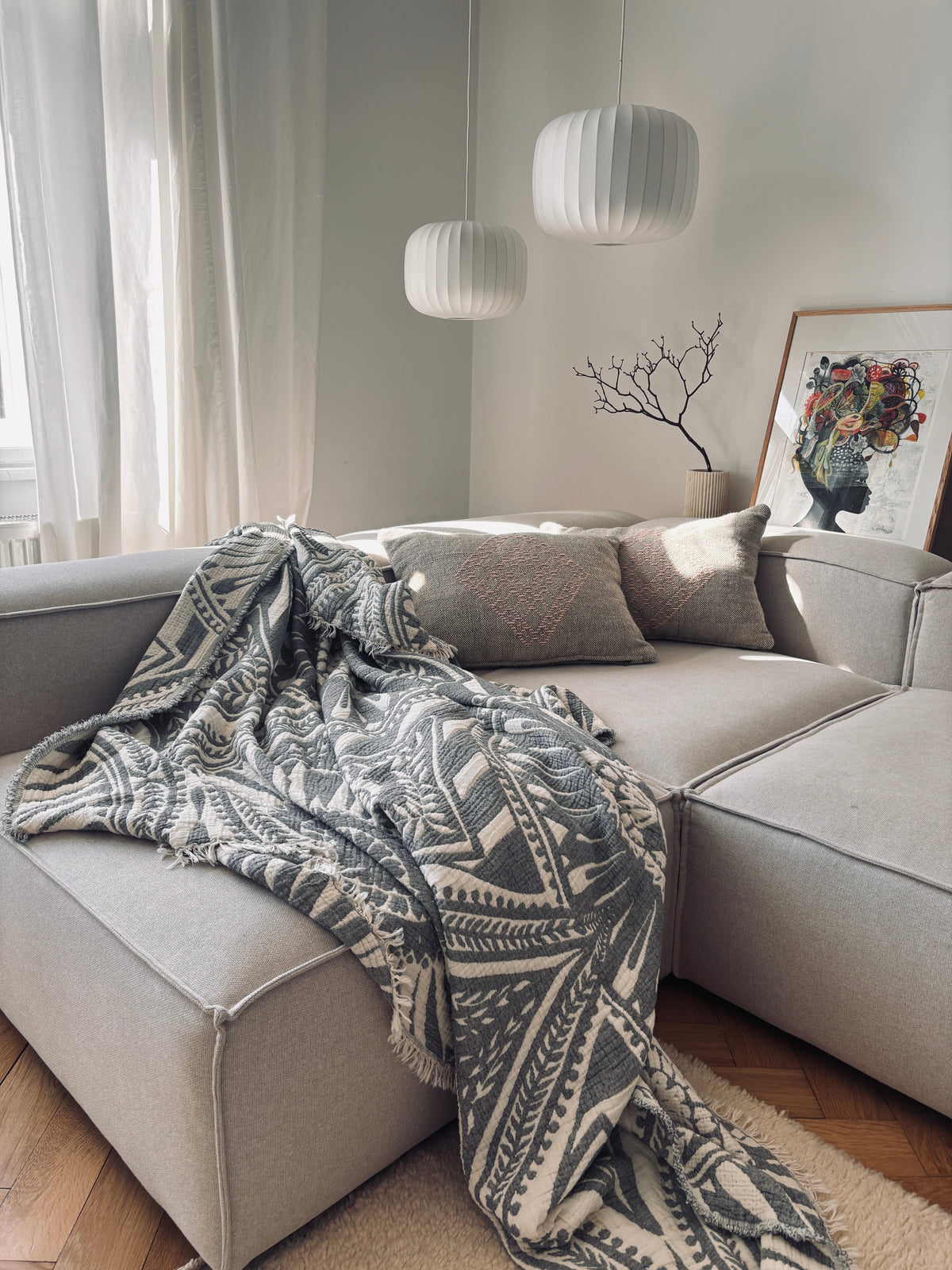 Ein gemütliches Wohnzimmer mit einem hellgrauen Sektionssofa, der LEY! CALMA Musselin Decke in Dunkelgrau (2,20 x 2,30 m), zwei strukturierte Kissen, Pendelleuchten, ein gerahmtes abstraktes Porträt, ein dekorativer Ast und transparente Vorhänge mit natürlichem Licht.