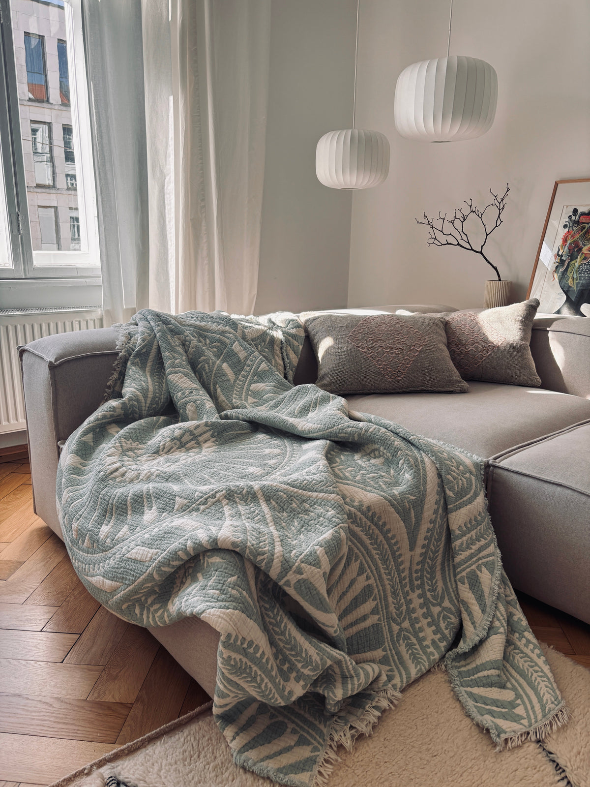 Ein gemütliches Wohnzimmer mit einem grauen Sektionssofa, drapiert mit der LEY! CALMA Musselin Decke in Mint (2,20 x 2,30 m), dekorativen Kissen, weißen Pendelleuchten, gerahmten Kunstwerken und einem Fenster, das natürliches Licht hereinlässt.
