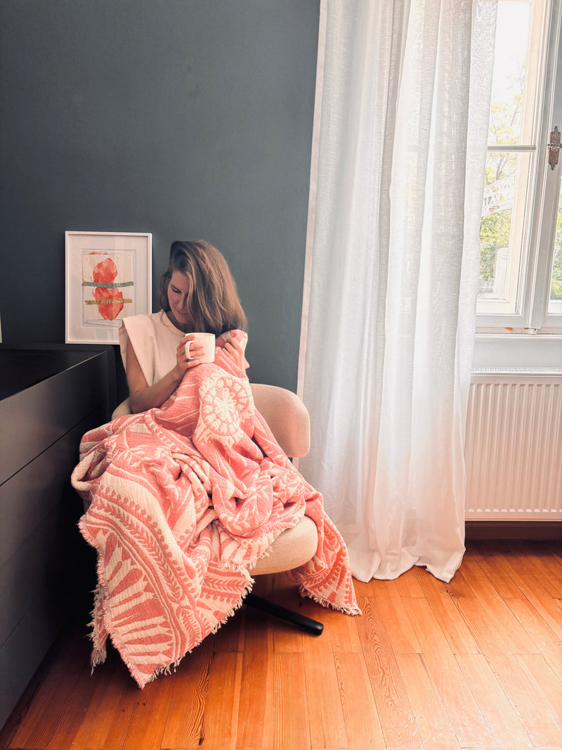 Frau am Fenster in eine CALMA Musselin Decke - Koralle von LEY! gehüllt, Sonnenlicht fällt ein.