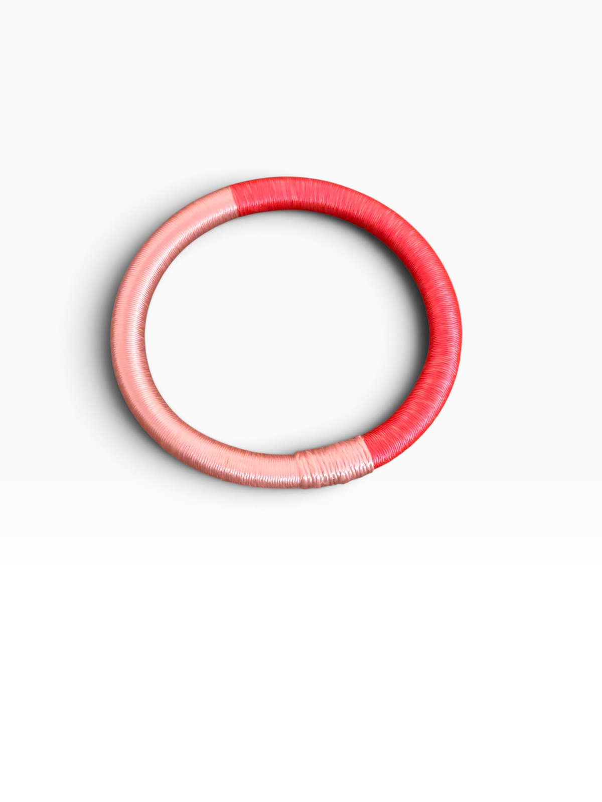 Ein runder Armreif „Minama – Koralle-Pink“ von LEY!, umwickelt mit rosa und rotem Faden, liegt auf einem strahlend weißen Hintergrund.