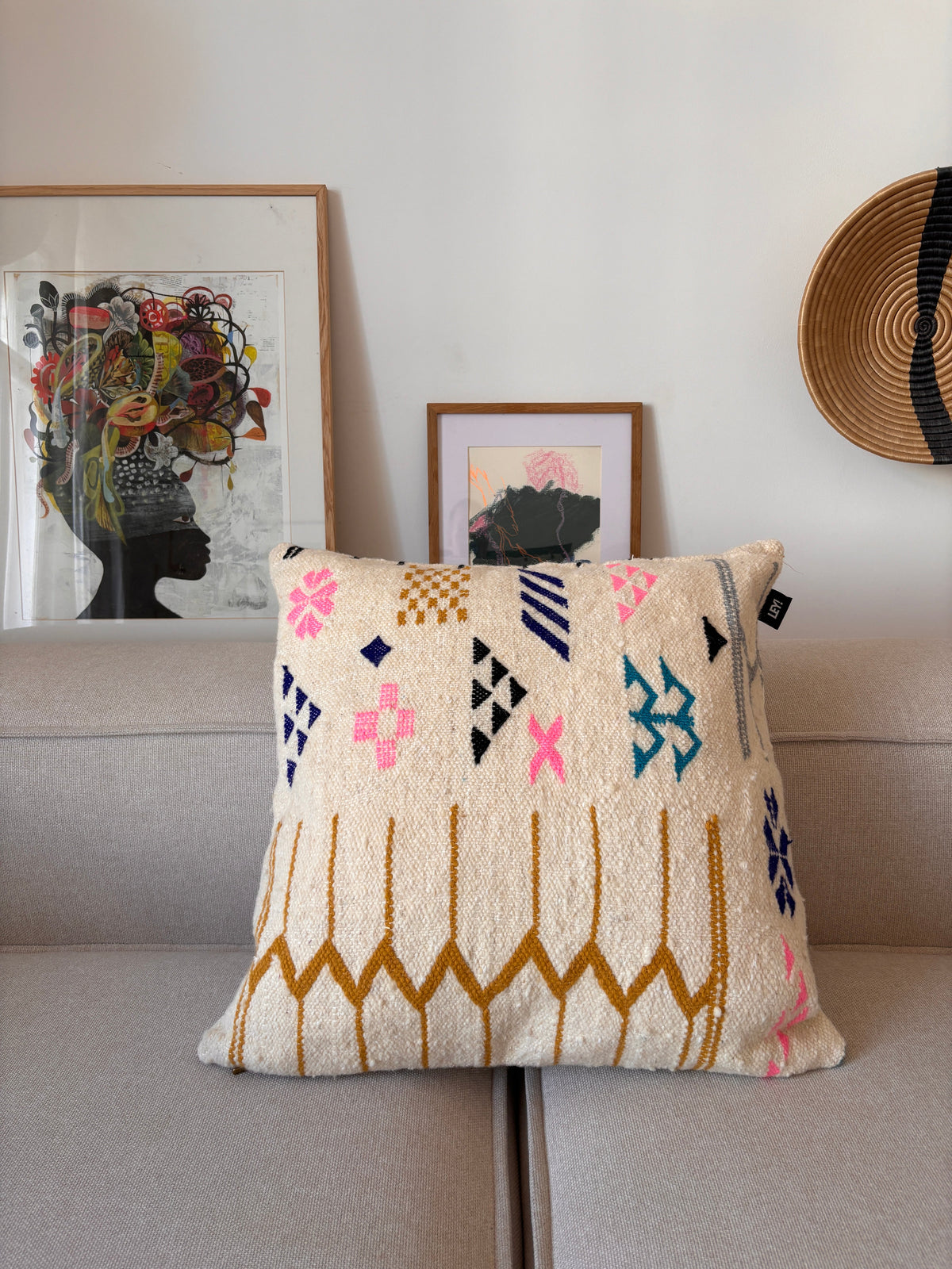 EIN LEY! Kissen - Colorful Line (60x60) von LEY! sitzt auf einem grauen Sofa, gerahmte Kunst und ein geflochtener Hut hängen an der weißen Wand.