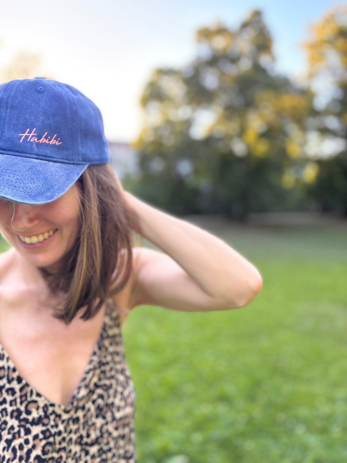 Eine lächelnde Frau in LEY! Mit einer „Habibi“-Kappe in Blau und einem Kleid mit Leopardenmuster steht sie auf einer Wiese.