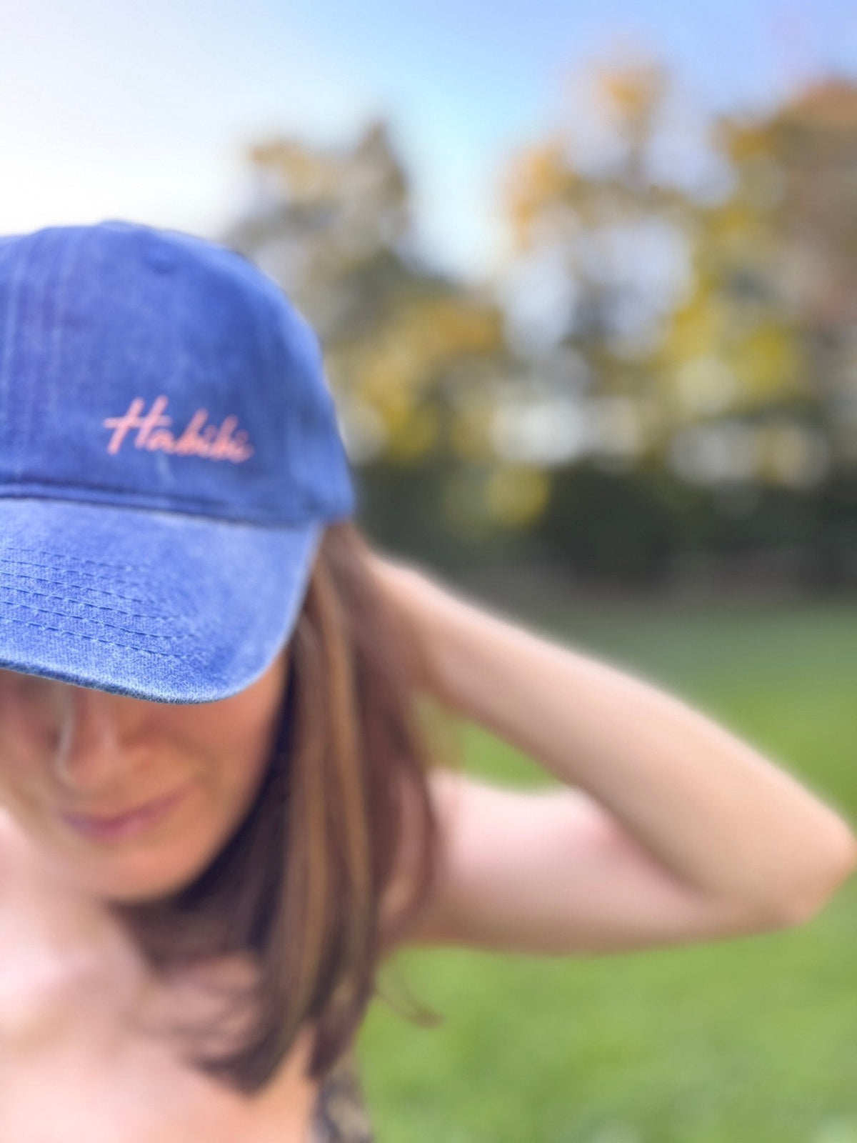 Eine Frau trägt eine LEY! „Habibi“-Kappe in Blau mit verstellbarem Verschluss und steht im Freien zwischen verschwommenen Bäumen und Gras.