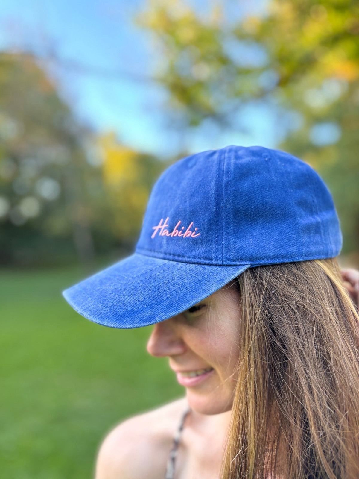 Eine Frau im Freien mit einer „Habibi“-Kappe von LEY! in Blau, die vor einem verschwommenen grünen Hintergrund lächelt.