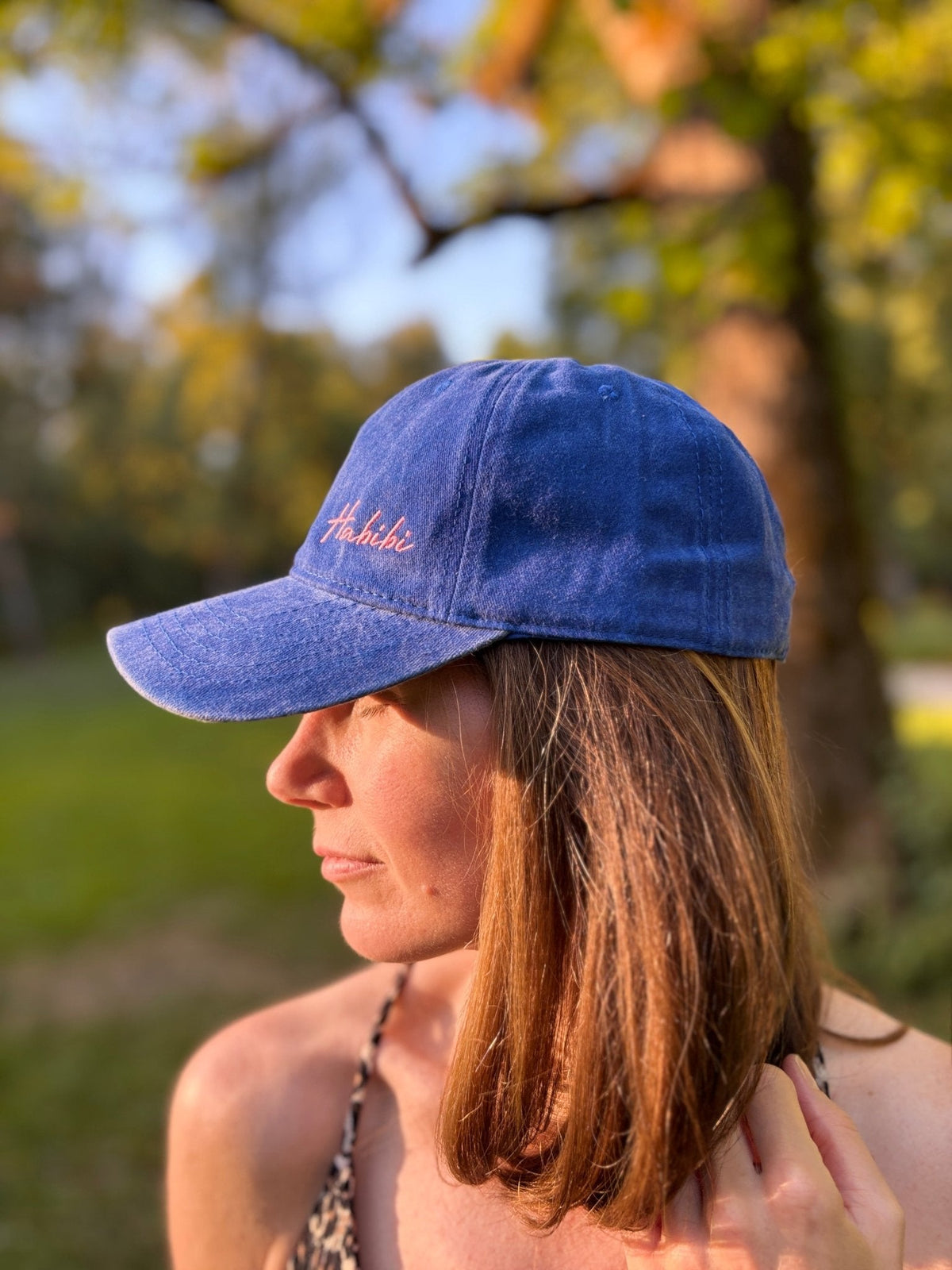 Eine Frau trägt die LEY! „Habibi“-Kappe in Blau im Freien, Sonnenlicht auf ihrem Gesicht und verschwommene Bäume im Hintergrund.