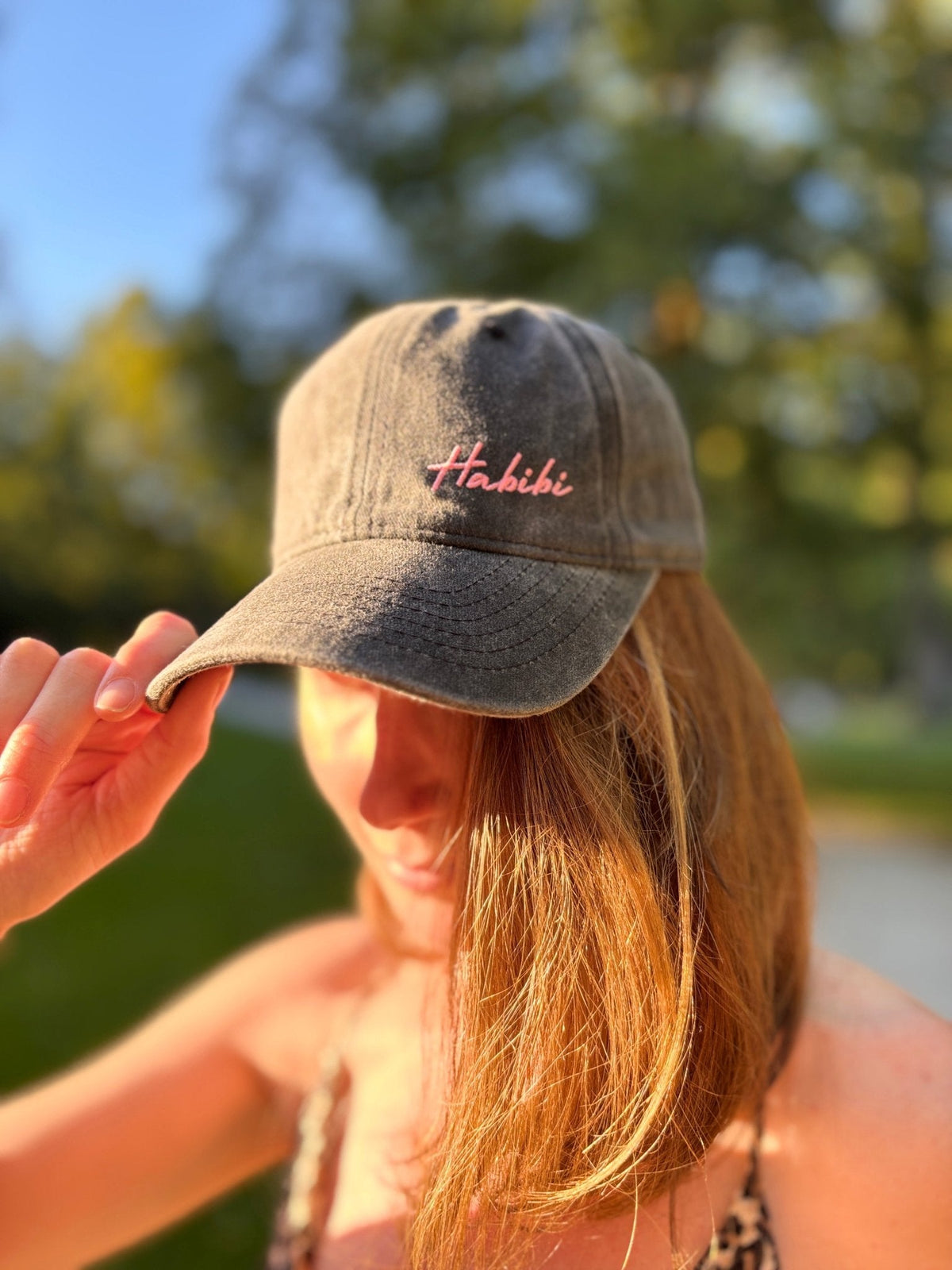 Eine Frau im Sonnenlicht trägt die LEY! „Habibi“-Kappe in Rosa mit verstellbarem Riemen, die die Krempe berührt; der Hintergrund ist unscharf.