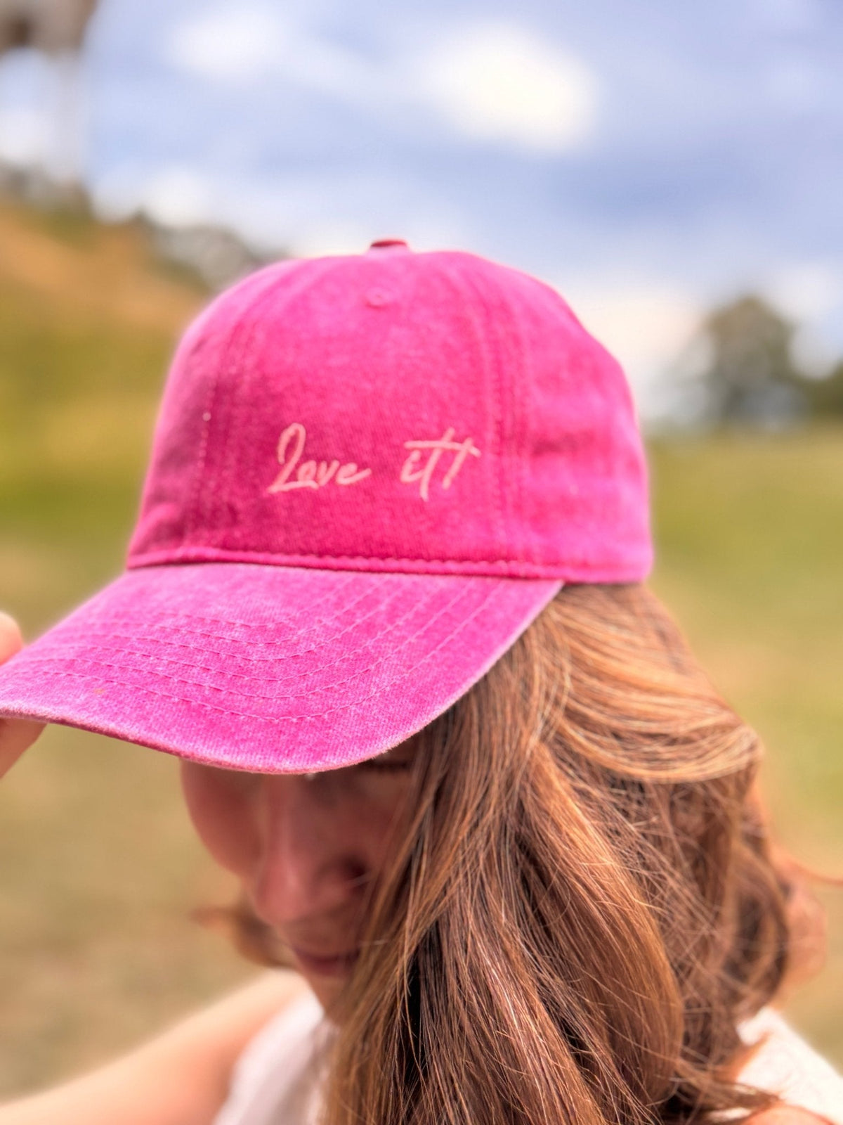 Eine Frau mit gewelltem Haar trägt draußen die „Love it“-Kappe in Rosa von LEY! und kreiert damit einen stilvollen Unisex-Look.