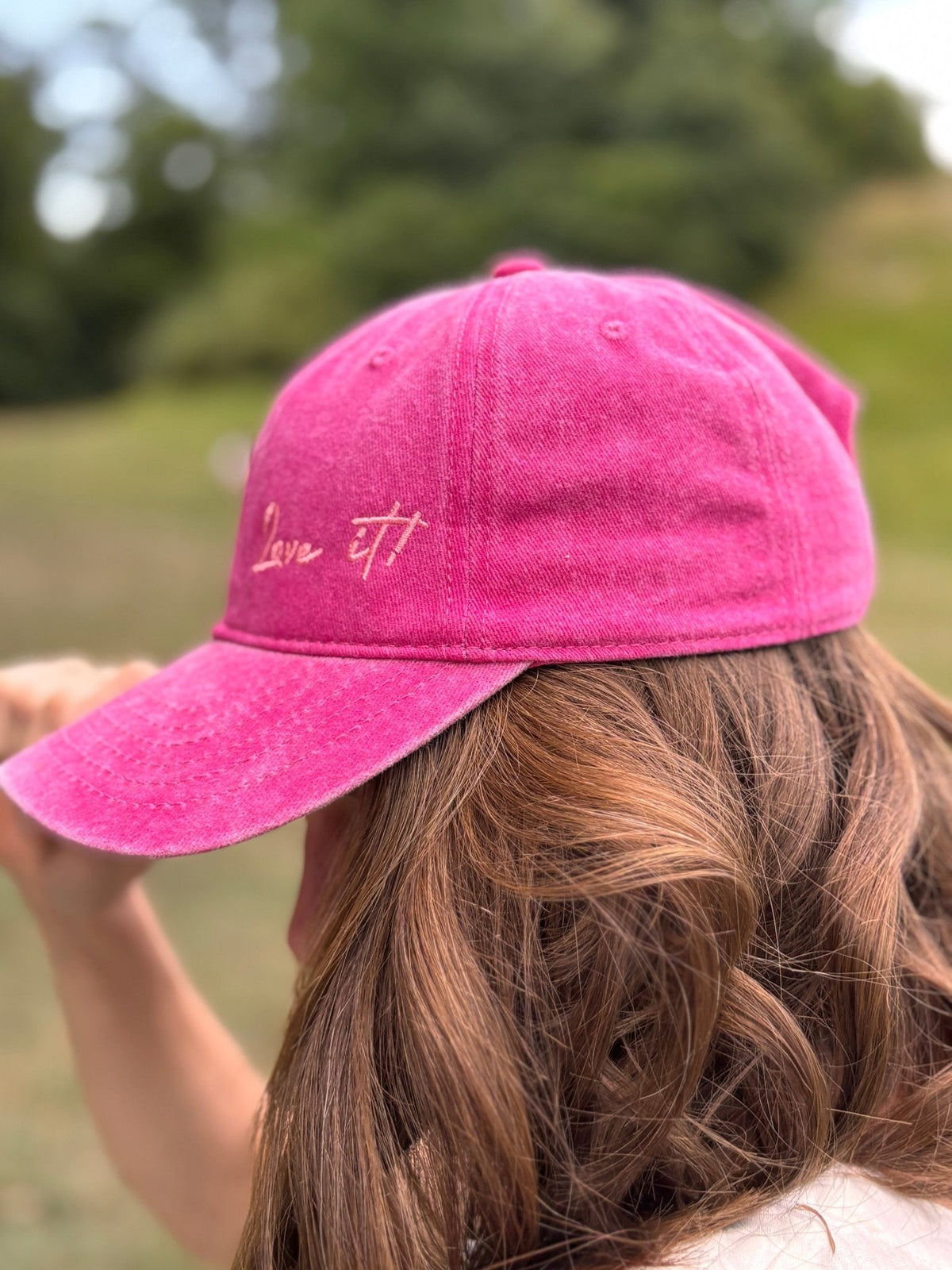 Eine Person mit welligem braunem Haar, die draußen eine grüne LEY!-Kappe mit der Aufschrift „Love it“ trägt, im Hintergrund ist Grün zu sehen.
