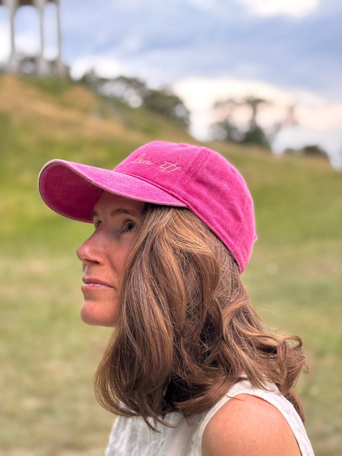 Eine Frau mit welligem braunem Haar trägt die LEY! „Love it“-Kappe in Rosa und ein weißes Oberteil, draußen vor dem Hintergrund von Gras und Bäumen.
