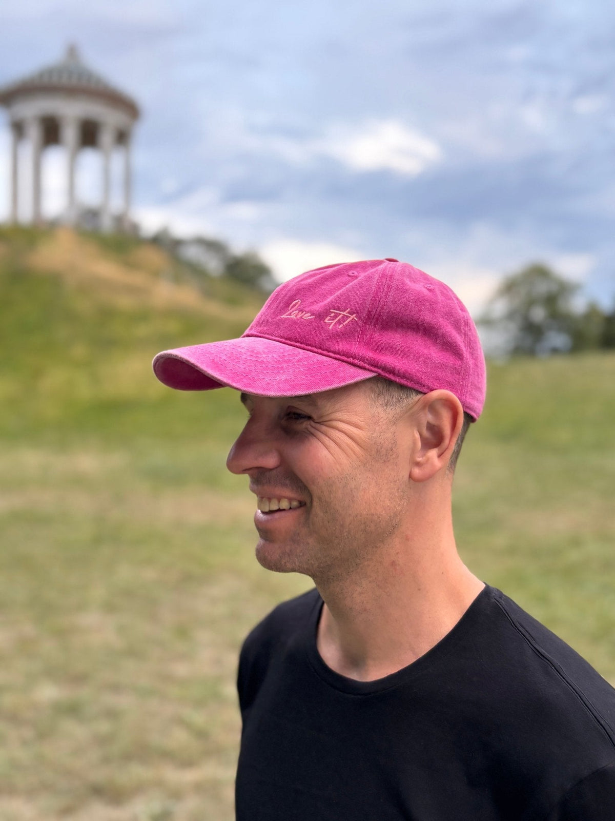 Ein Mann mit einer LEY!-Kappe mit der Aufschrift „Love it“ und einem rosa-schwarzen T-Shirt lächelt im Freien vor einem grasbewachsenen Hügel und einem Pavillon.
