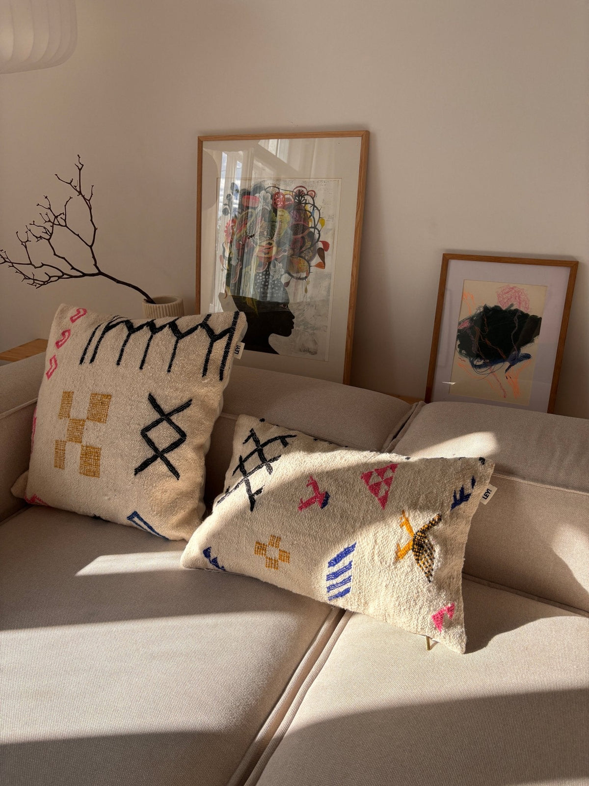 Zwei LEY!-Kissen – „Colorful Line“ 40 x 60 auf einem beigen Sofa, Sonnenstrahlen, die hereinscheinen, gerahmte Kunstwerke und ein Ast im Hintergrund.
