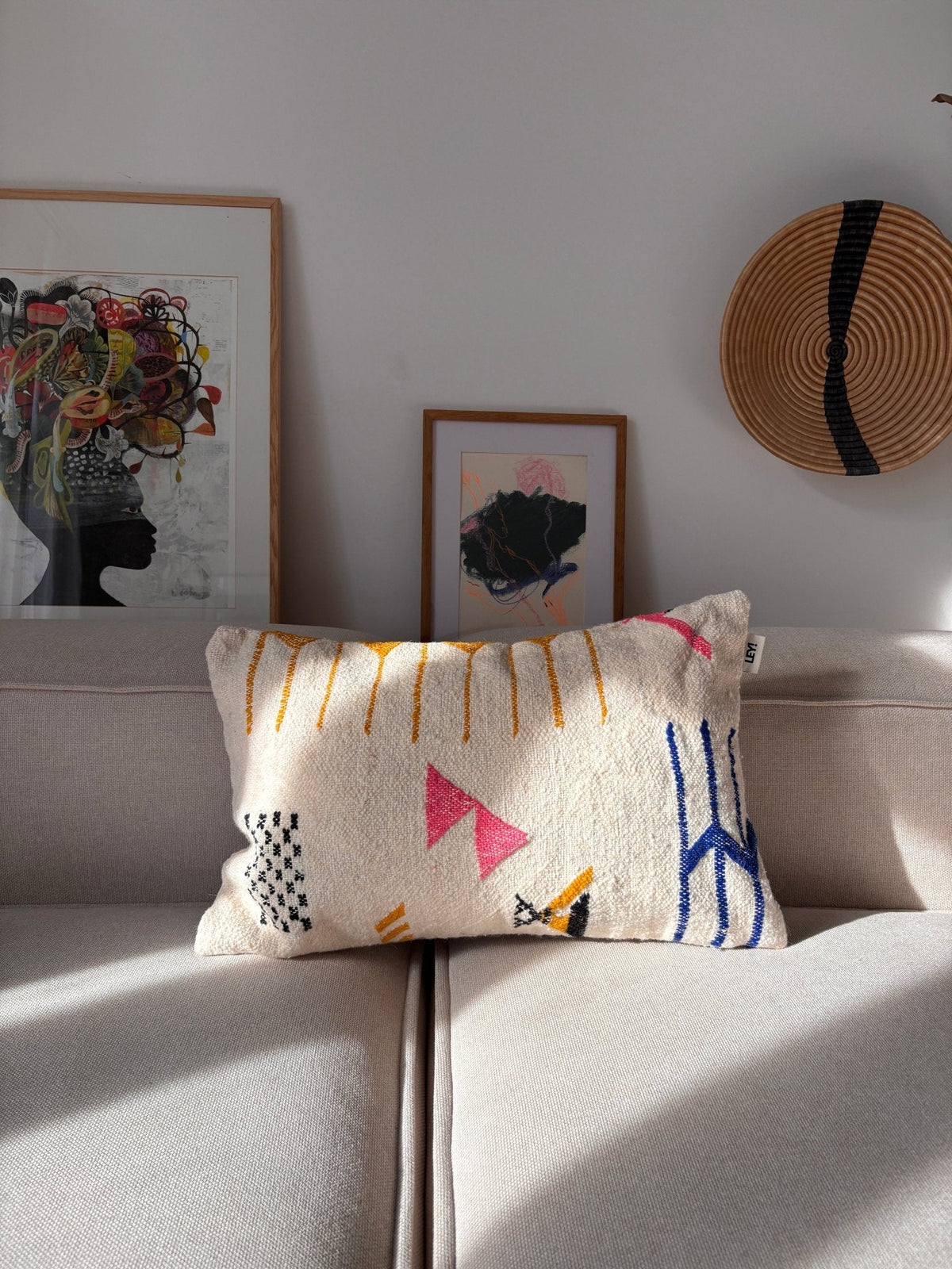 Beiges Sofa mit einem „LEY!“-Kissen – „Colorful Line“ – 40 x 60, gerahmtes Kunstwerk und geflochtener Korb an einer sonnenbeschienenen weißen Wand.