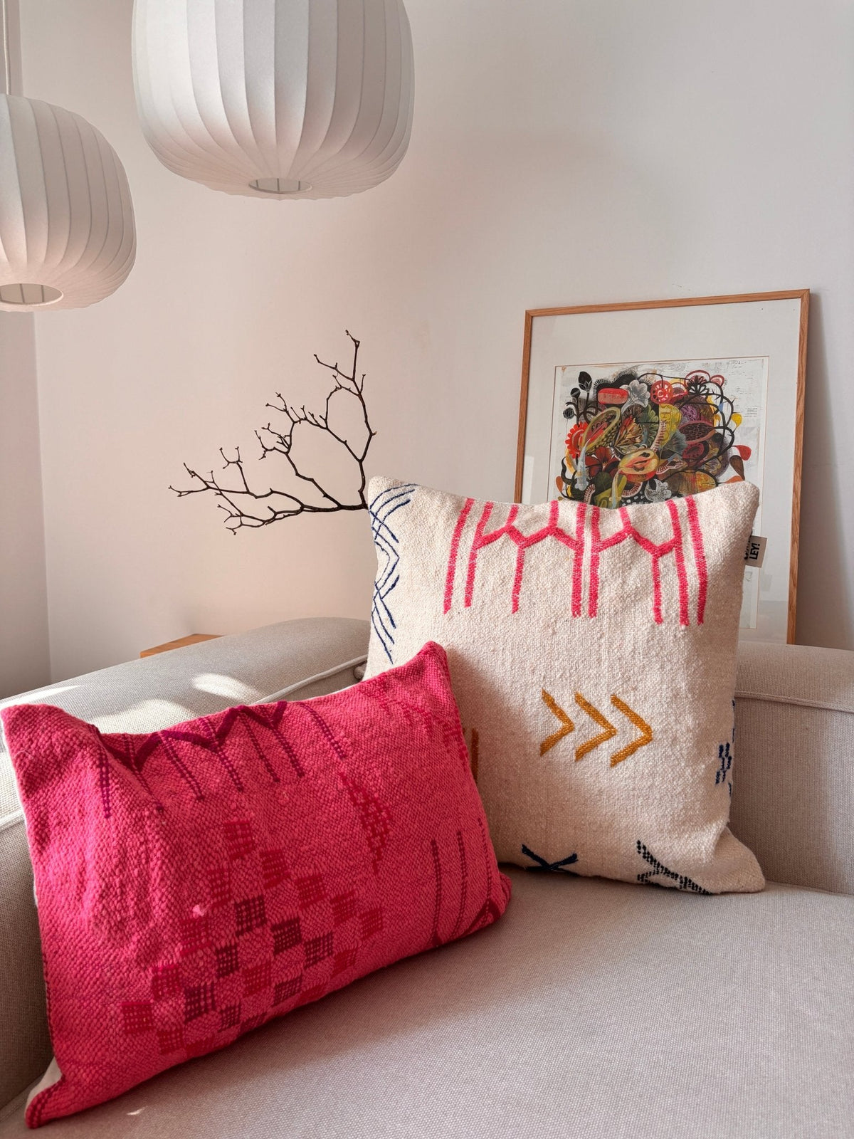 Zwei LEY!-Kissen – „Colorful Line“, 60 x 60 cm, auf einem beigen Sofa in einem gemütlichen, sonnendurchfluteten Raum mit Lampen und gerahmten Kunstwerken.