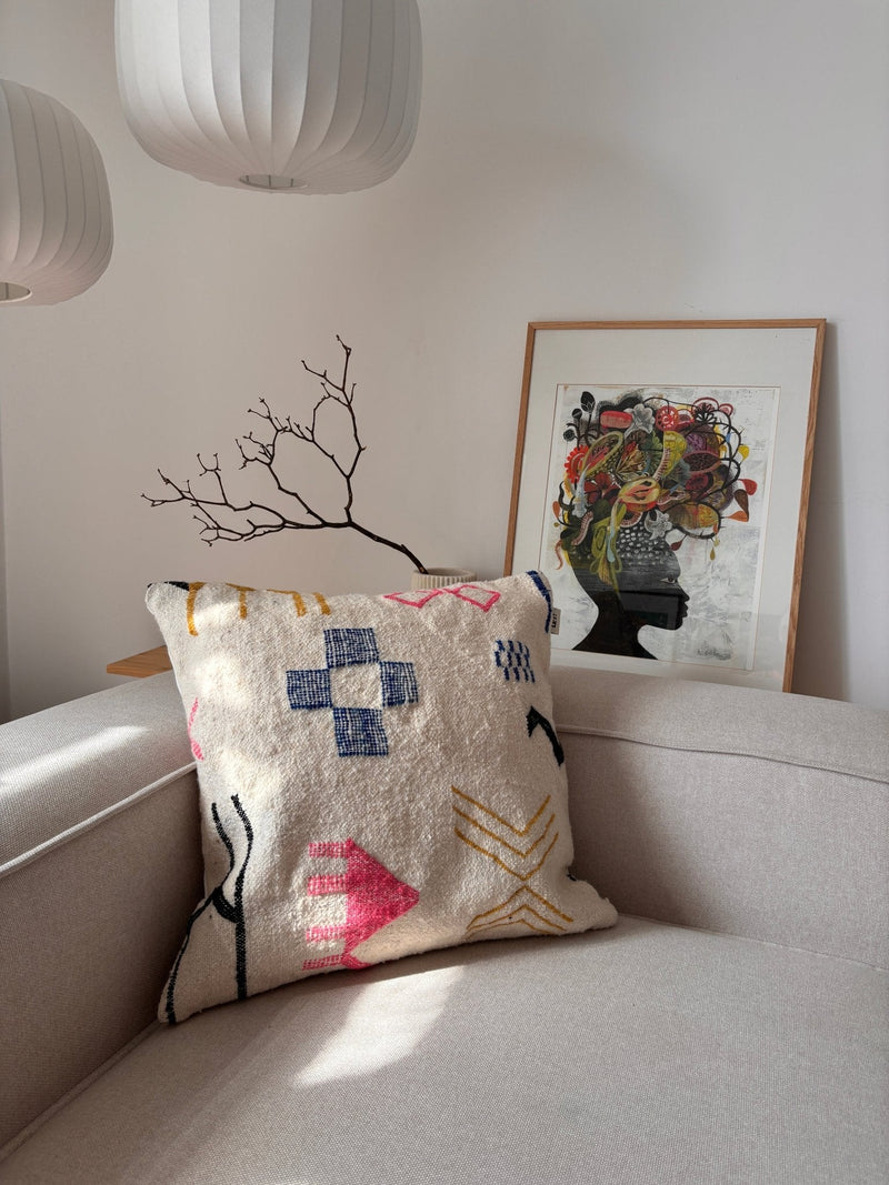 Ein LEY!-Kissen – „Colorful Line“, 60 x 60, auf einem beigen Sofa, kombiniert mit Kunstwerken und Lampen in einem hellen, stilvollen Raum.