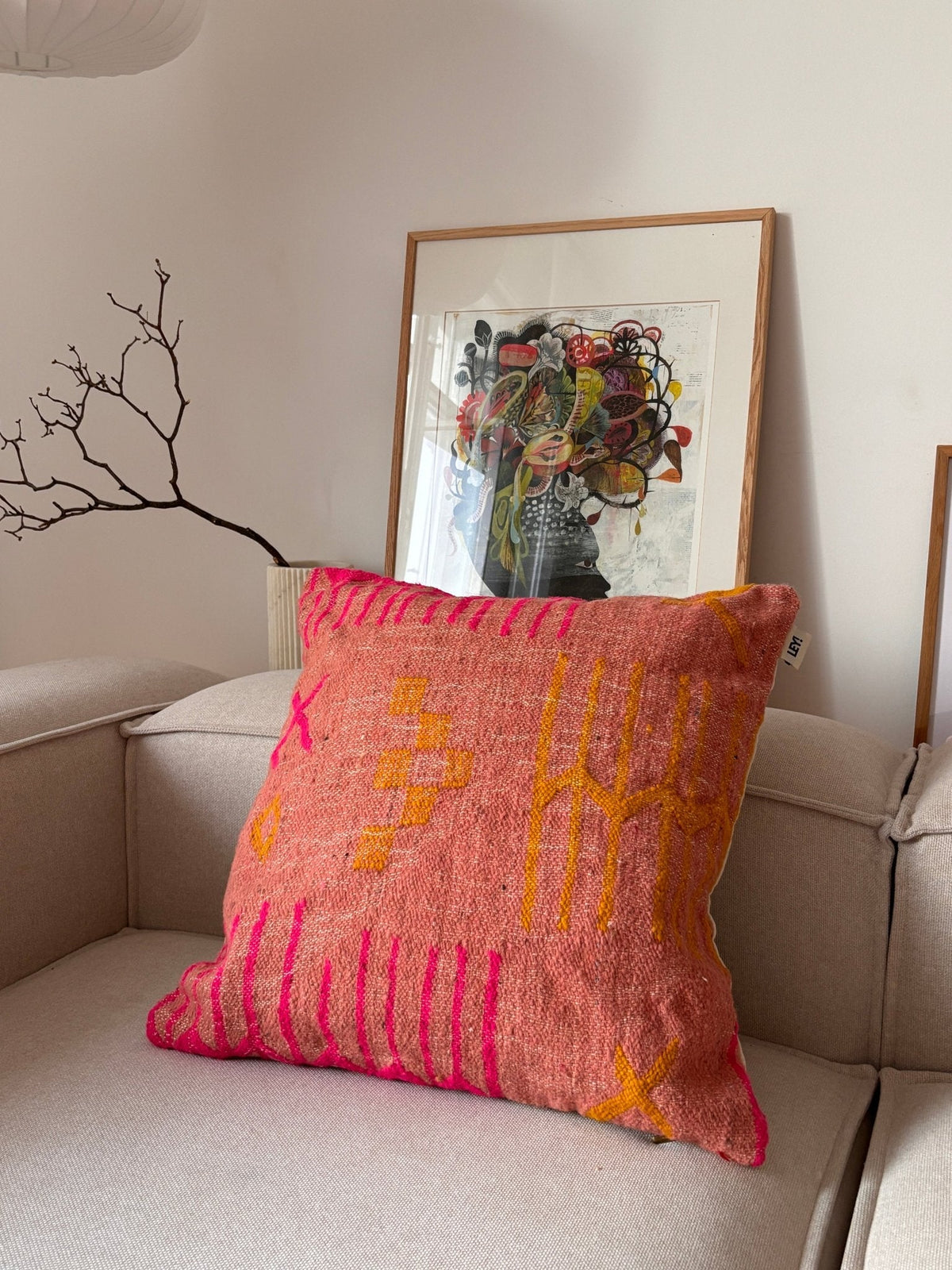 Ein LEY!-Kissen – Colorful Line – Terracotta Edition verleiht einem beigen Sofa, einem gerahmten Kunstwerk und einem kahlen Ast einen modernen Touch.