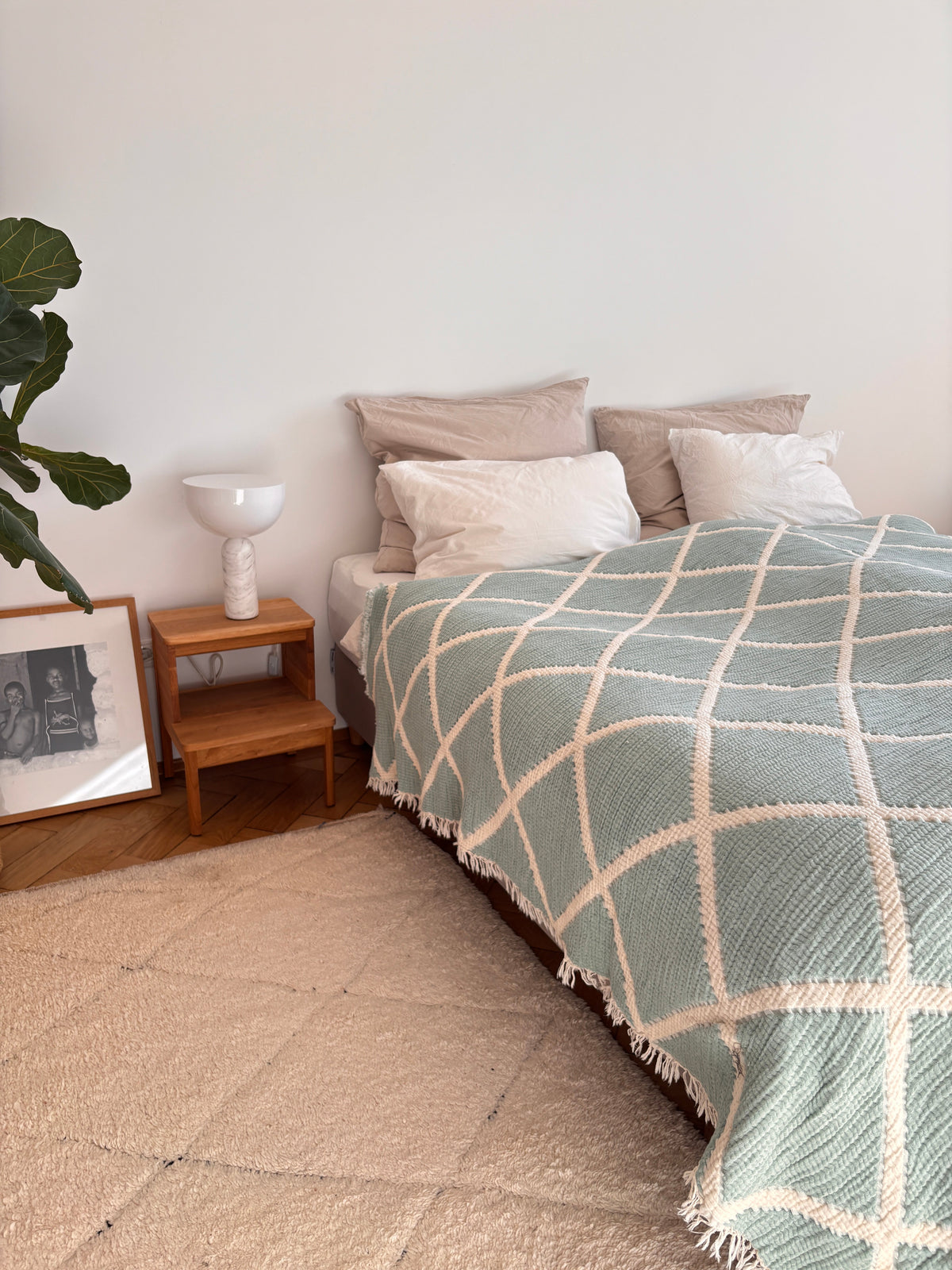 Ein ordentlich gemachtes Bett mit beigen und weißen Kissen wird mit der LEY! Musselin Decke in Grau-Weiß (2,20 x 2,50 m), die für Boho-Flair sorgt. Auf einem hölzernen Nachttisch steht eine weiße Lampe, daneben stehen eine Blattpflanze und ein gerahmtes Schwarz-Weiß-Foto.