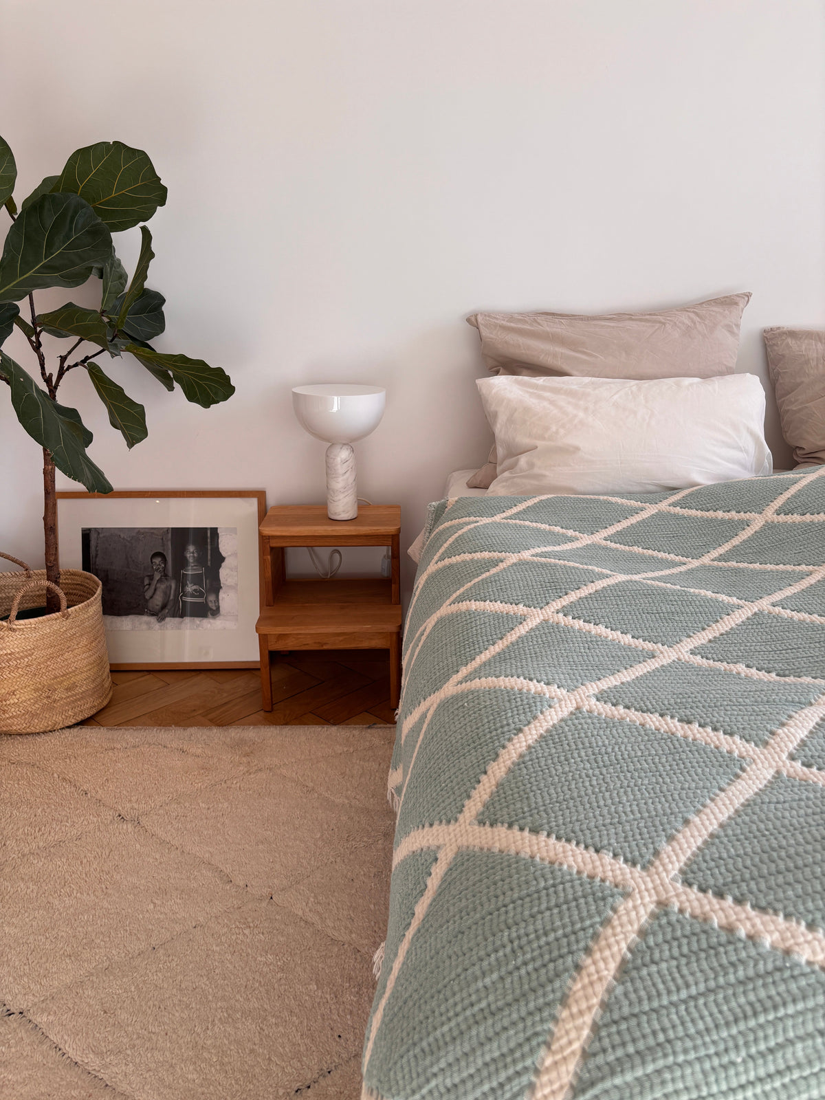 Eine gemütliche Schlafzimmerecke mit der LEY! Musselin Decke in Grau-Weiß (2,20 x 2,50 m) auf dem Bett, einem kleinen Holznachttisch mit weißer Lampe und Vase, einem gerahmten Foto auf dem Boden, einer großen Topfpflanze und einem beigen Teppich.