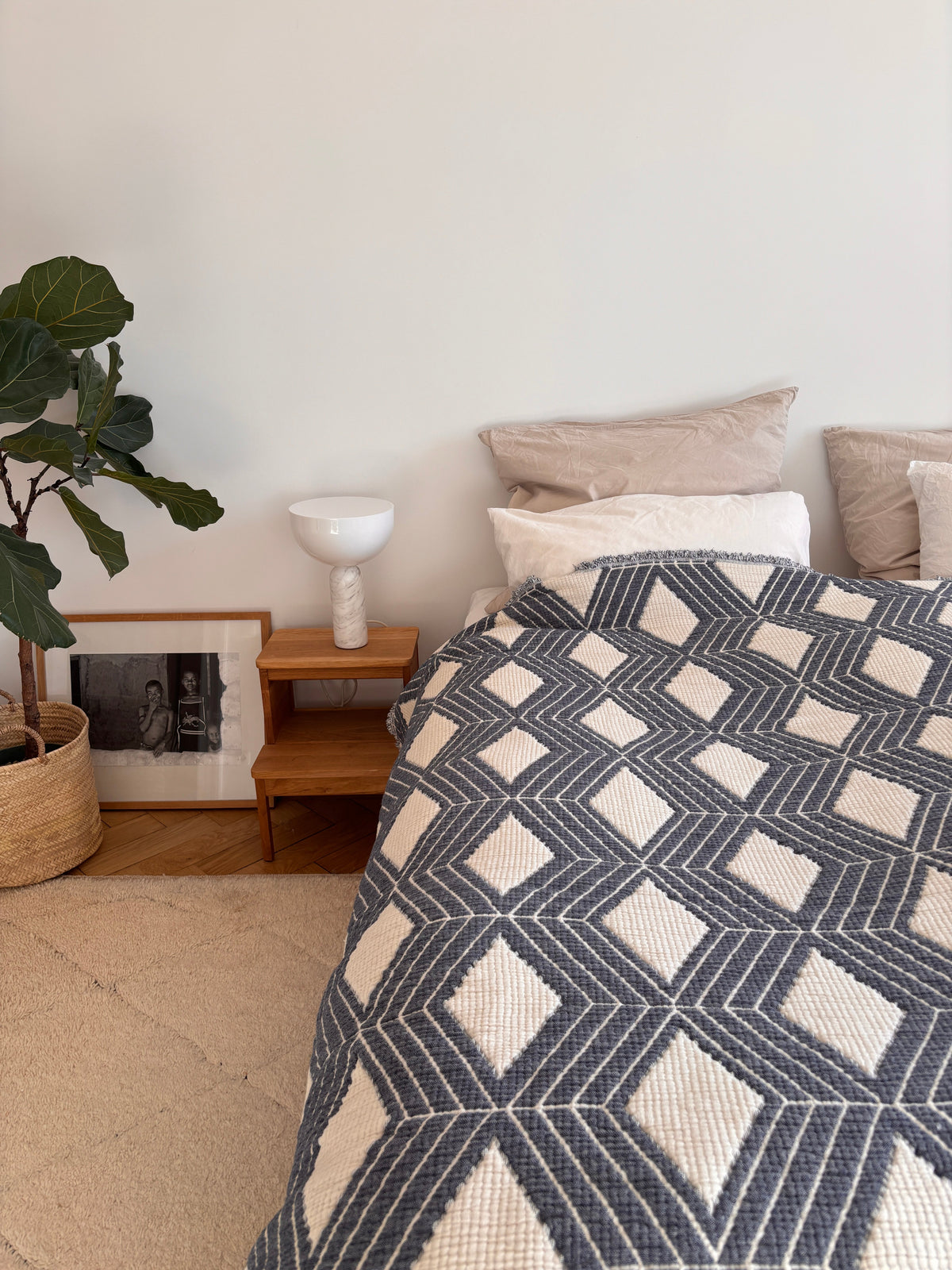 Ein modernes Schlafzimmer mit einem Hauch von Boho-Design, mit einem Bett, das mit der LEY! Musselin Decke - Blau-Weiß (2,20 x 2,50 m), beigen Kissen, einem hölzernen Nachttisch mit einer weißen Lampe, einer Topfpflanze und einem gerahmten Foto an der Wand.