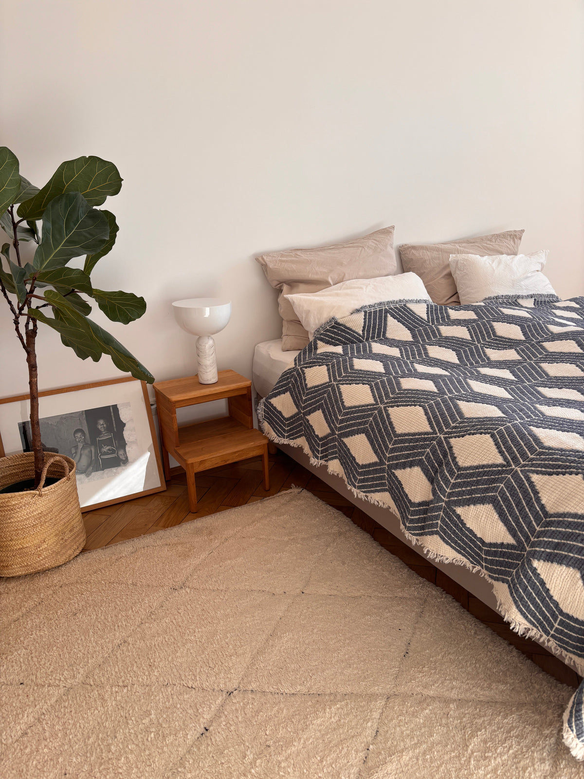 Ein gemütliches Schlafzimmer mit einem Bett, bezogen mit der LEY! Musselin Decke - Blau-Weiß (2,20 x 2,50 m), beigen Kissen, einem hölzernen Nachttisch mit weißer Lampe, Topfpflanzen und gerahmten Kunstwerken auf dem Boden für Boho-Design-Flair.