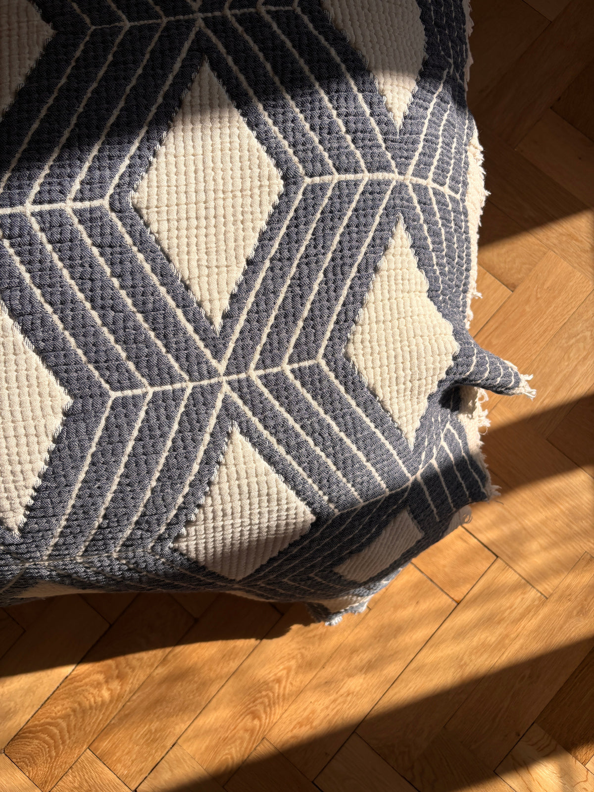 Ein blau-weißer, geometrischer Teppich liegt auf einem Holzboden, dessen Boho-Akzente durch das Sonnenlicht besonders schön zur Geltung kommen - eine schöne Ergänzung zur LEY! Musselin Decke in Blau-Weiß (2,20 x 2,50 m) für einen gemütlichen und stilvollen Look.