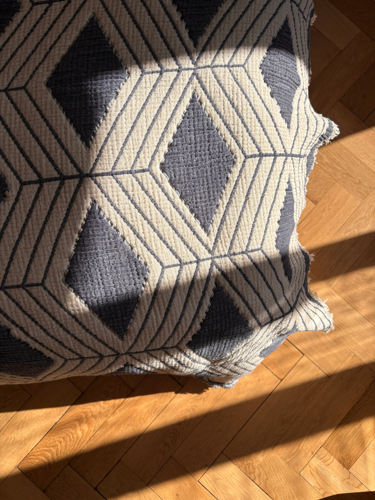 Eine Nahaufnahme der LEY! Musselin Decke in Blau-Weiß (2,20 x 2,50 m) liegt auf einem Holzboden. Das Sonnenlicht wirft diagonale Schatten, die die weiche Textur und das subtile Boho-Design betonen.