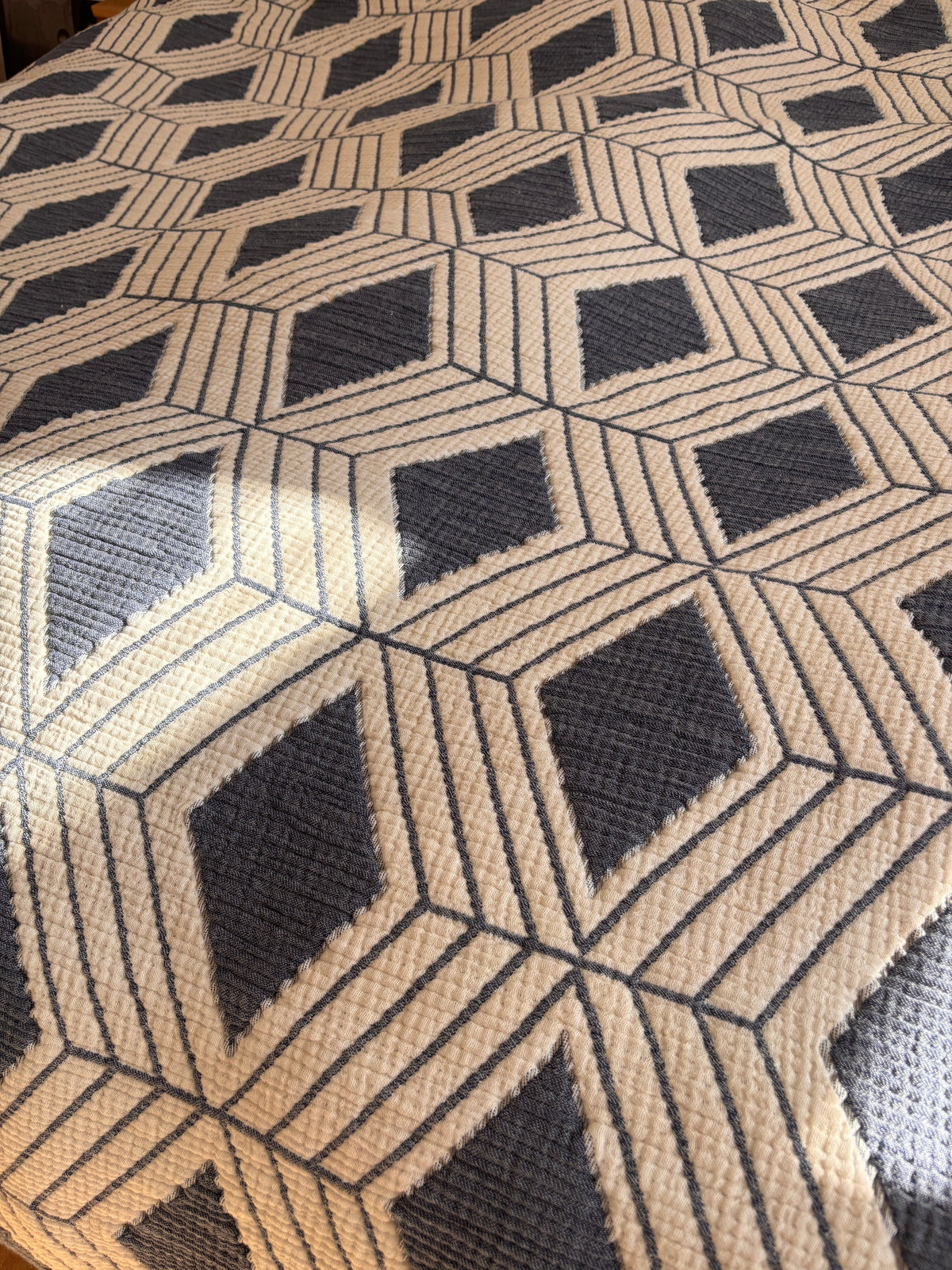 Die Nahaufnahme der Musselin Decke - Blau-Weiß (2,20 x 2,50 m) von LEY! zeigt ein geometrisches Boho-Muster in Marine und Creme mit Rautenformen und sonnenbeschienenen Linien - stilvoll und perfekt für Ihren Raum.