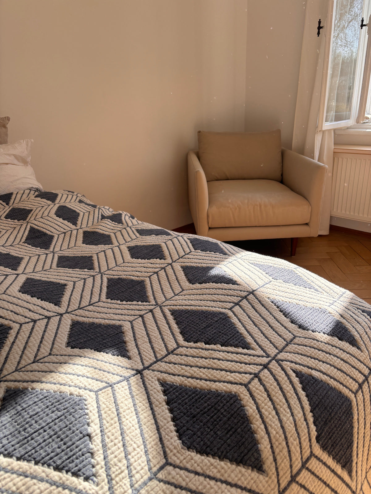 Ein sonnendurchflutetes Schlafzimmer mit der LEY! Musselin Decke in Blau-Weiß (2,20 x 2,50 m) mit geometrischem Boho-Design, einem beigen Sessel am Fenster und Sonnenlicht, das auf den Parkettboden fällt.