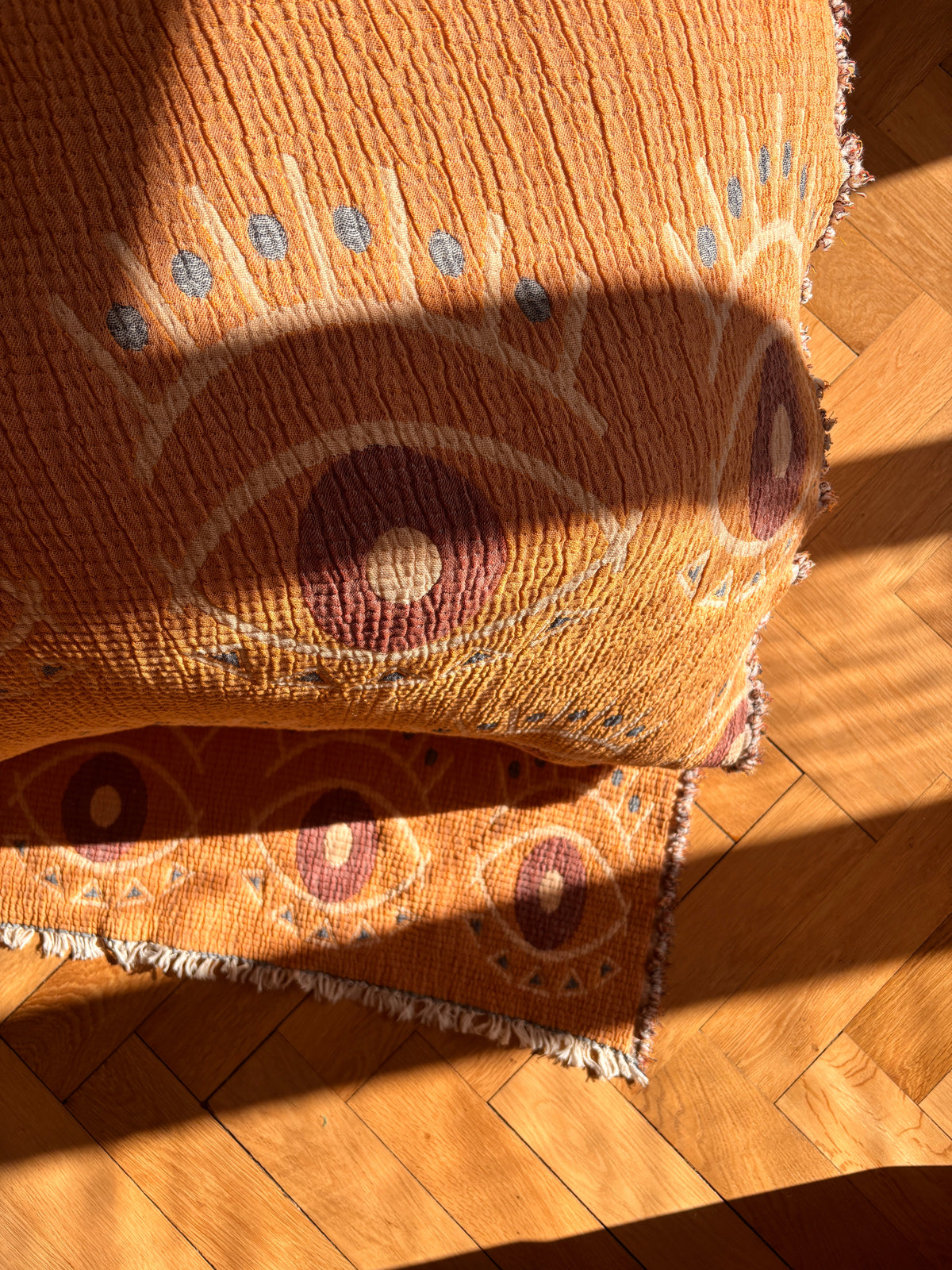 Die Musselin Decke - Vision - Gelb-Blau (2,20 x 2,50 m) von LEY! verleiht dem sonnenbeschienenen, orangefarbenen Teppich mit Augenmuster und weißen Fransen auf einem Parkettboden eine gemütliche Textur, während Sonnenlicht und Schatten klare Linien über die Szene ziehen.