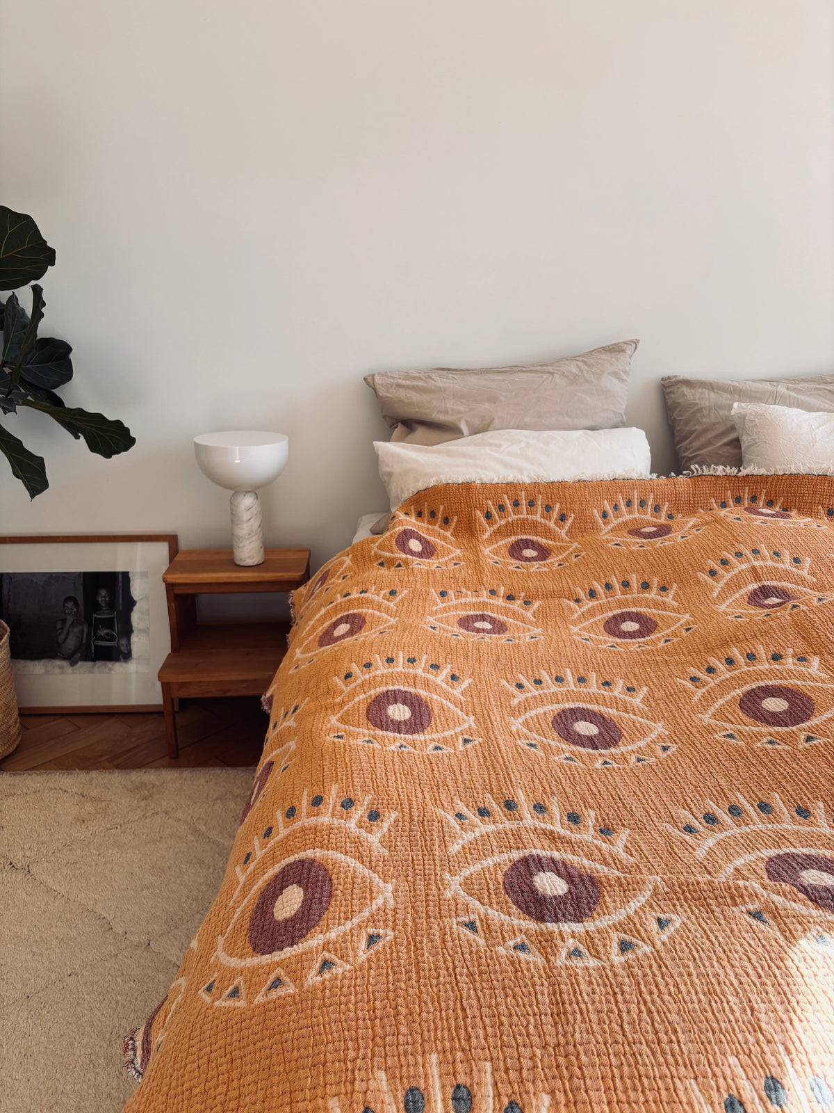 Ein gemütliches Bett mit beigen Kissen und einer LEY! Musselin Decke - Vision in gelb-blau (2,20 x 2,50 m) sorgt für Gemütlichkeit. Ein kleiner Holznachttisch mit einer weißen Lampe und einem gerahmten Foto steht neben einer grünen Topfpflanze.